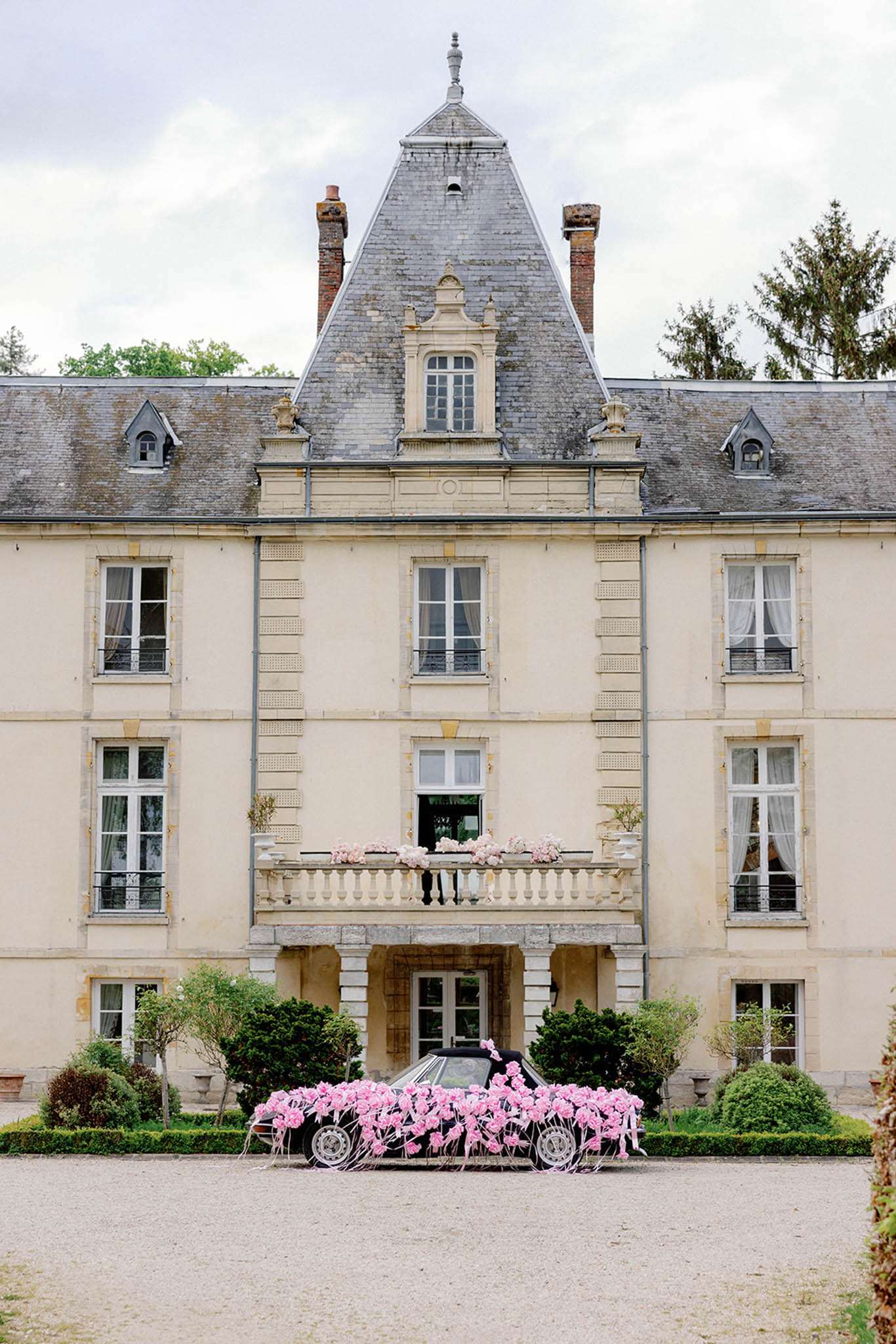 A wide shot of the front facade of a French château, featuring a cream-colored stone exterior with a central pointed slate roof tower, symmetrical windows, and a first-floor balustrade balcony decorated with pink floral arrangements. Parked on the gravel driveway directly in front of the entrance is a dark vintage car covered almost entirely in pink roses and tied with white ribbon streamers, serving as a decorated wedding getaway car. The balcony above the entrance also displays clusters of pink blooms, creating a coordinated pink floral theme across both the car and the building exterior. No people are visible in the frame. Potential venue feature image.