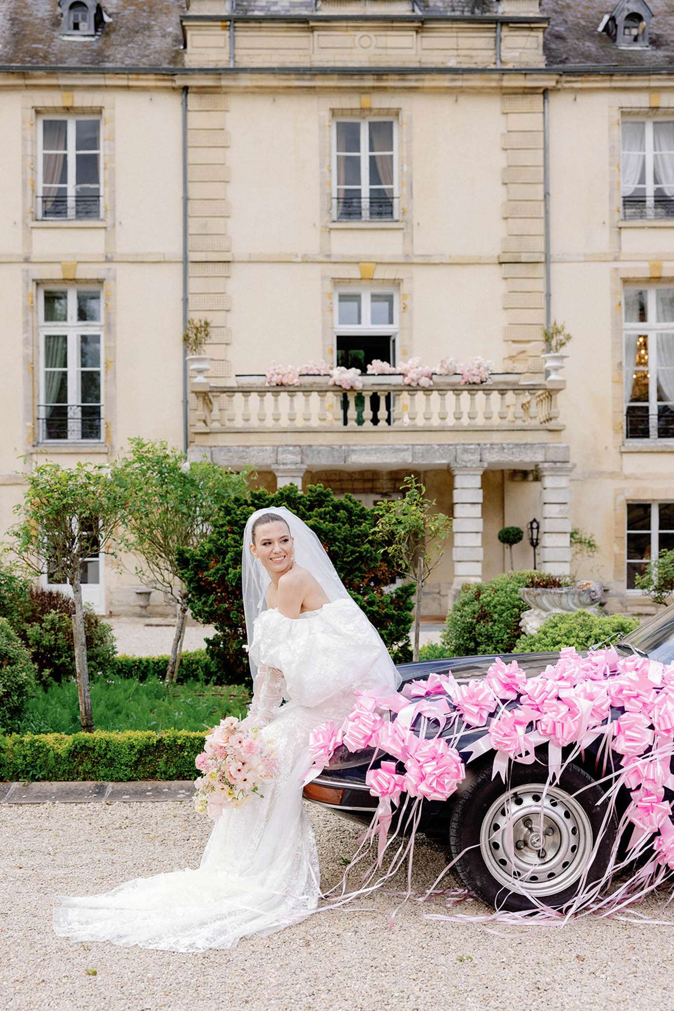 Bride in lace gown with cathedral veil beside vintage car with hot pink ribbons cream chateau with balcony florals behind