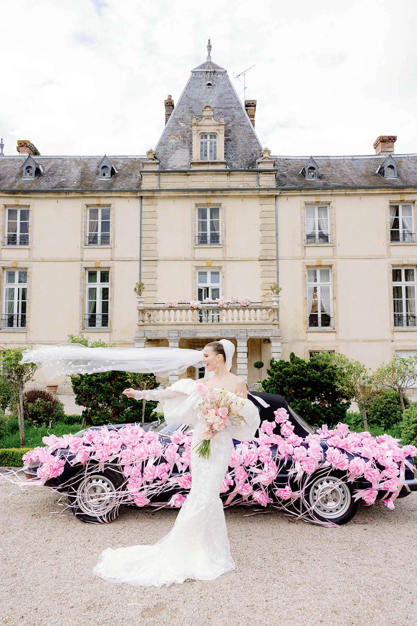 Bride in white lace gown with veil holding pink rose bouquet beside flower-decorated car at French chateau