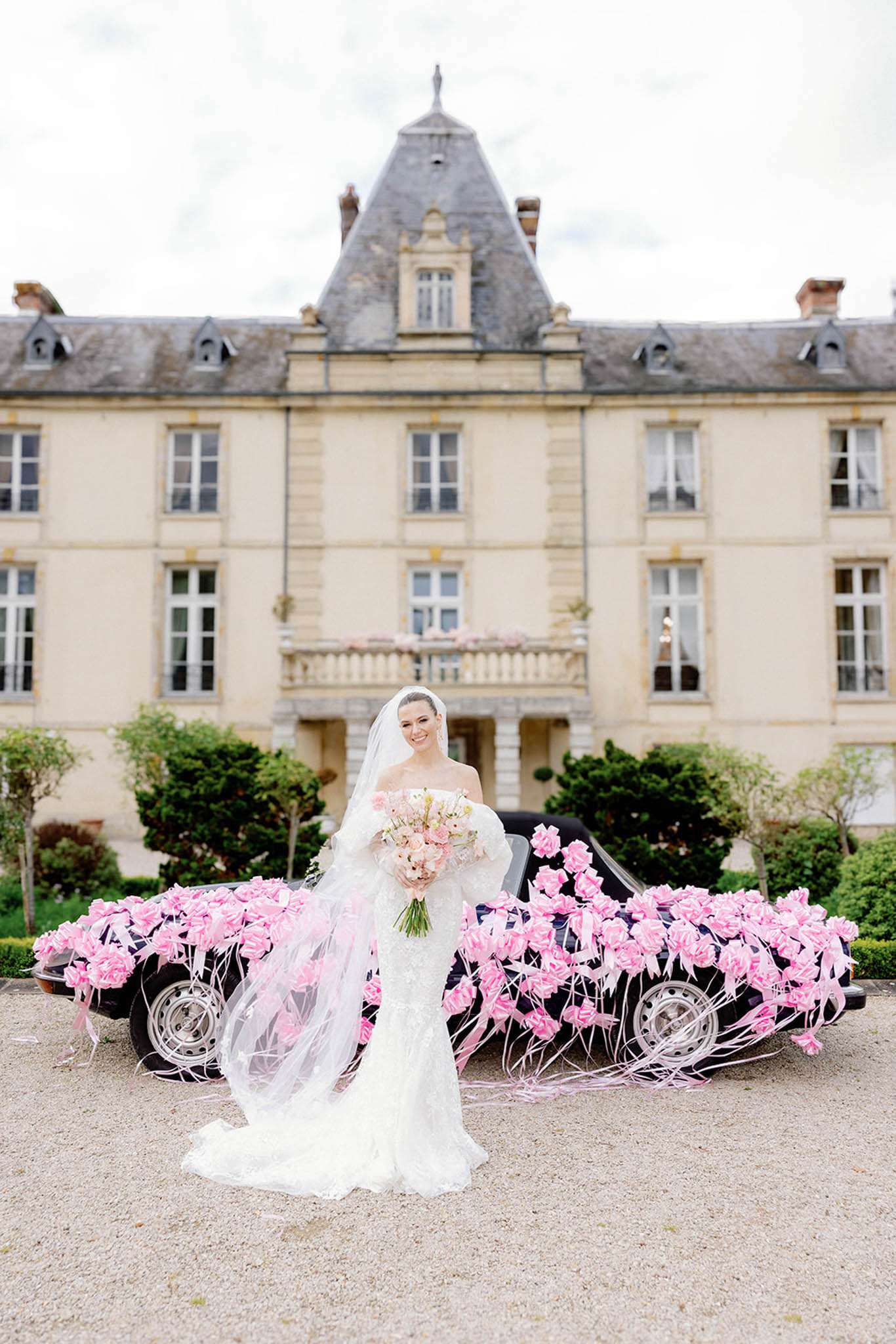 A bridal portrait taken outdoors on a gravel forecourt in front of a large French château with pale stone façade, slate-roofed turrets, and a central balcony. The bride stands alone wearing a white off-the-shoulder fitted lace gown with a cathedral-length train and a long cathedral veil, holding a bouquet of blush pink and ivory roses with trailing ribbon. Behind her, a dark vintage convertible car is densely decorated with bright pink paper or silk roses and pink ribbons covering the hood and sides entirely. The composition is a full-length portrait with the château centered symmetrically in the background. Potential venue feature image.