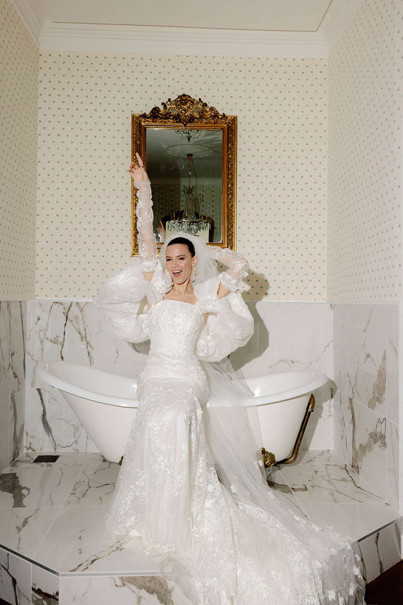 Bride laughing with arms raised in marble bathroom wearing fitted lace gown with cathedral veil and sheer sleeves