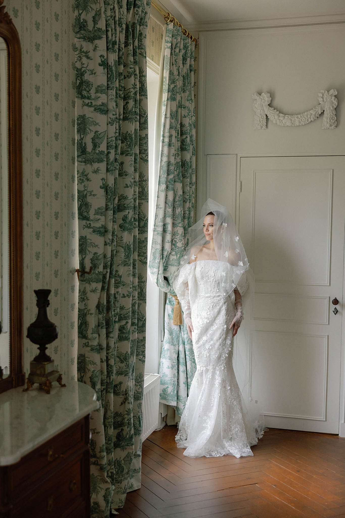 Bride in ivory lace mermaid gown and cathedral veil standing by window in chateau room with toile wallpaper