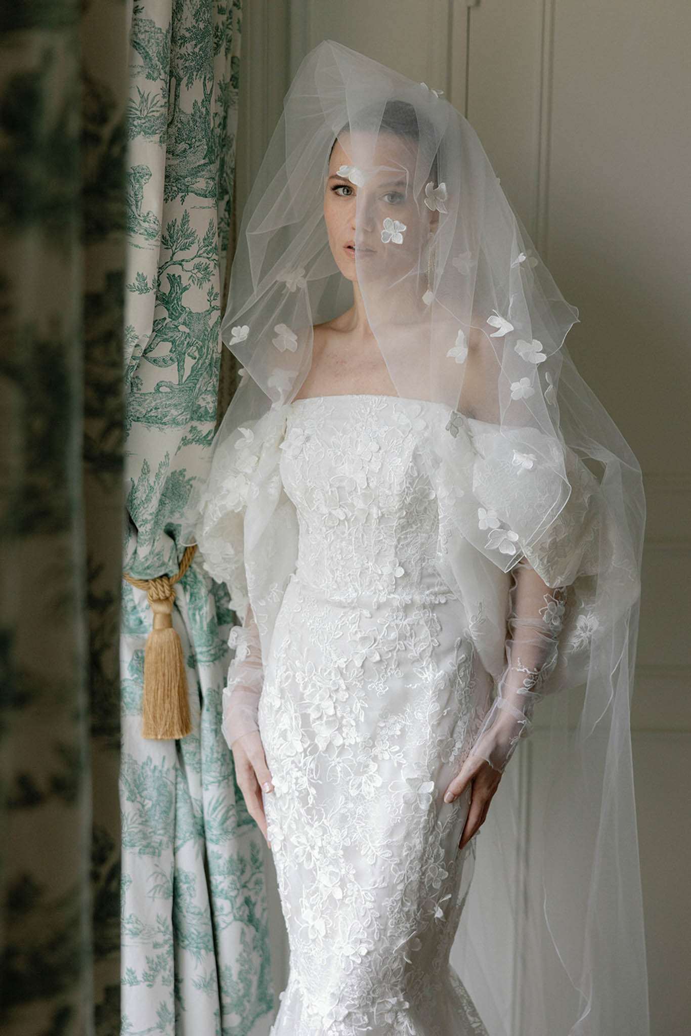 A bridal portrait of a single bride standing indoors beside a green and white toile de Jouy curtain tied back with a gold tassel. She wears a fitted, strapless white lace gown with all-over floral appliqué detailing and sheer long gloves with embroidered floral motifs. Her veil is draped forward over her face and features scattered white floral appliqués, matching the dress. The composition is a mid-length portrait shot with soft, diffused natural light. The classic French interior styling — panelled walls, toile fabric, tassel tie-back — contrasts with the sculptural, fashion-forward bridal look.