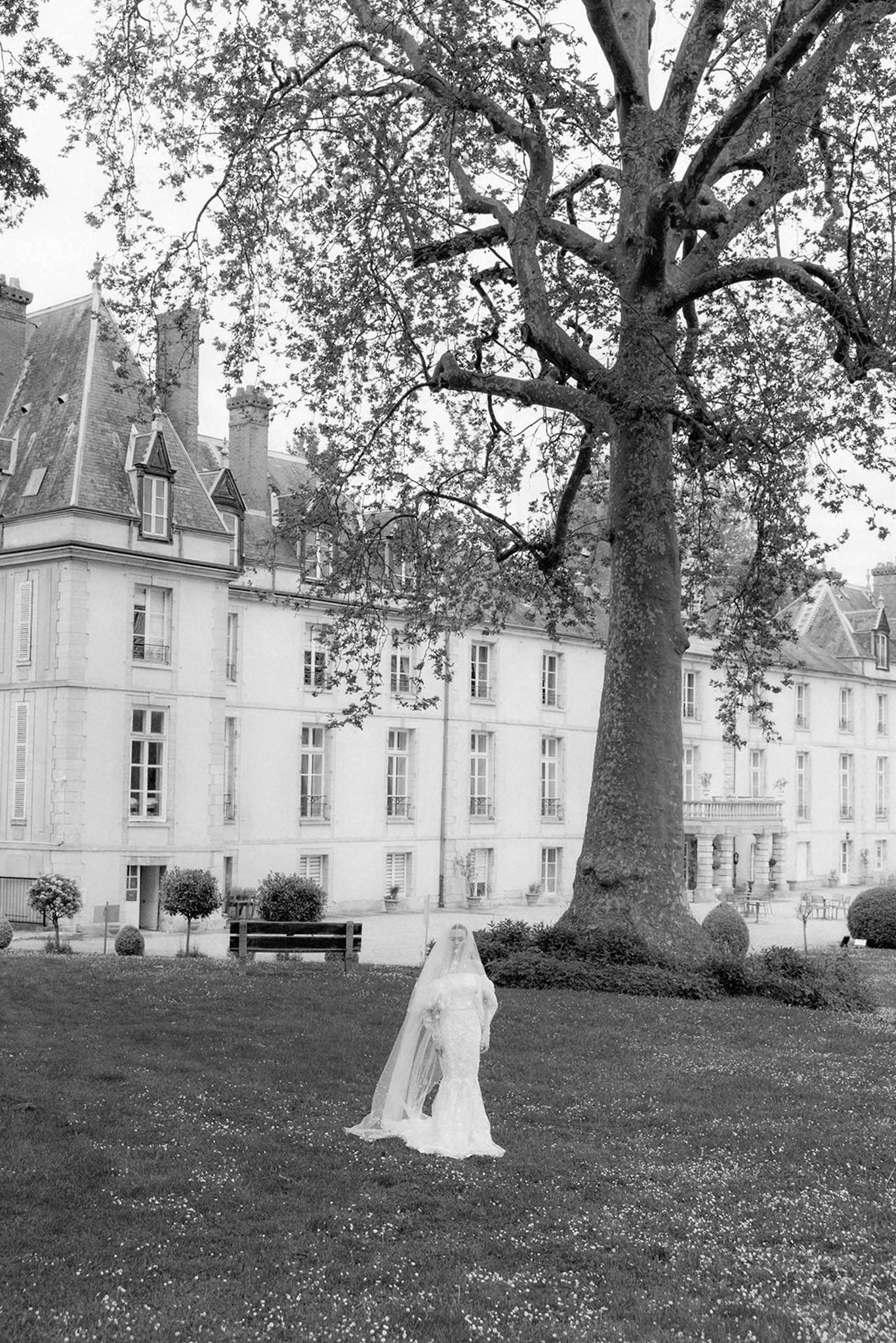 Black and white portrait of bride with cathedral veil standing on lawn before a French chateau