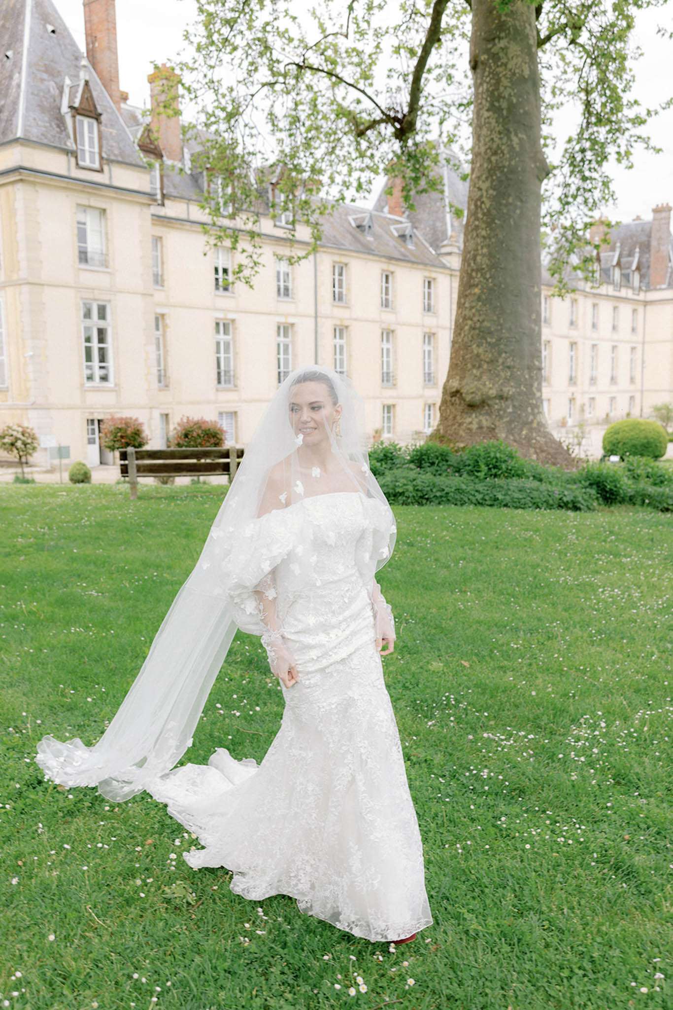 Bride in ivory lace mermaid gown with cathedral veil on chateau lawn smiling toward camera