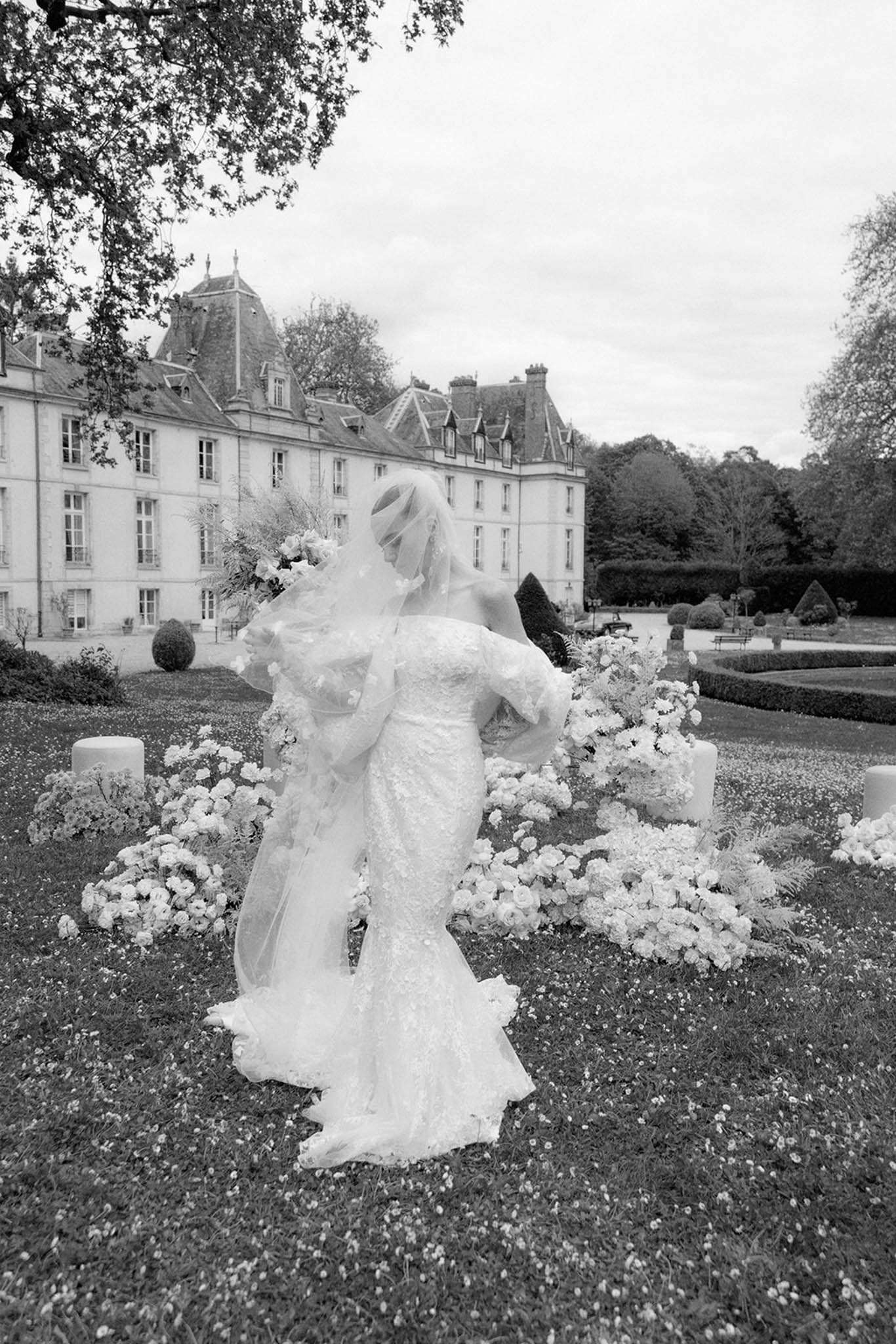 This black-and-white image captures a bride standing in the formal gardens of a large French château, whose grand multi-storey façade with mansard roofs is visible in the background. The bride wears a fitted lace gown with long sleeves and a long train, paired with a voluminous veil adorned with floral appliqués that billows around her in the wind, partially obscuring her face and figure. She is positioned among the ceremony floral arrangements, which consist of low garden-style clusters of full blooms — appearing to be roses and peonies — placed around tall white pillar candle stands on the lawn. The composition is a full-length portrait shot taken from slightly behind and to the side, with soft, even tones and moderate contrast that highlight the texture of the lace gown and the movement of the veil. Potential venue feature image.