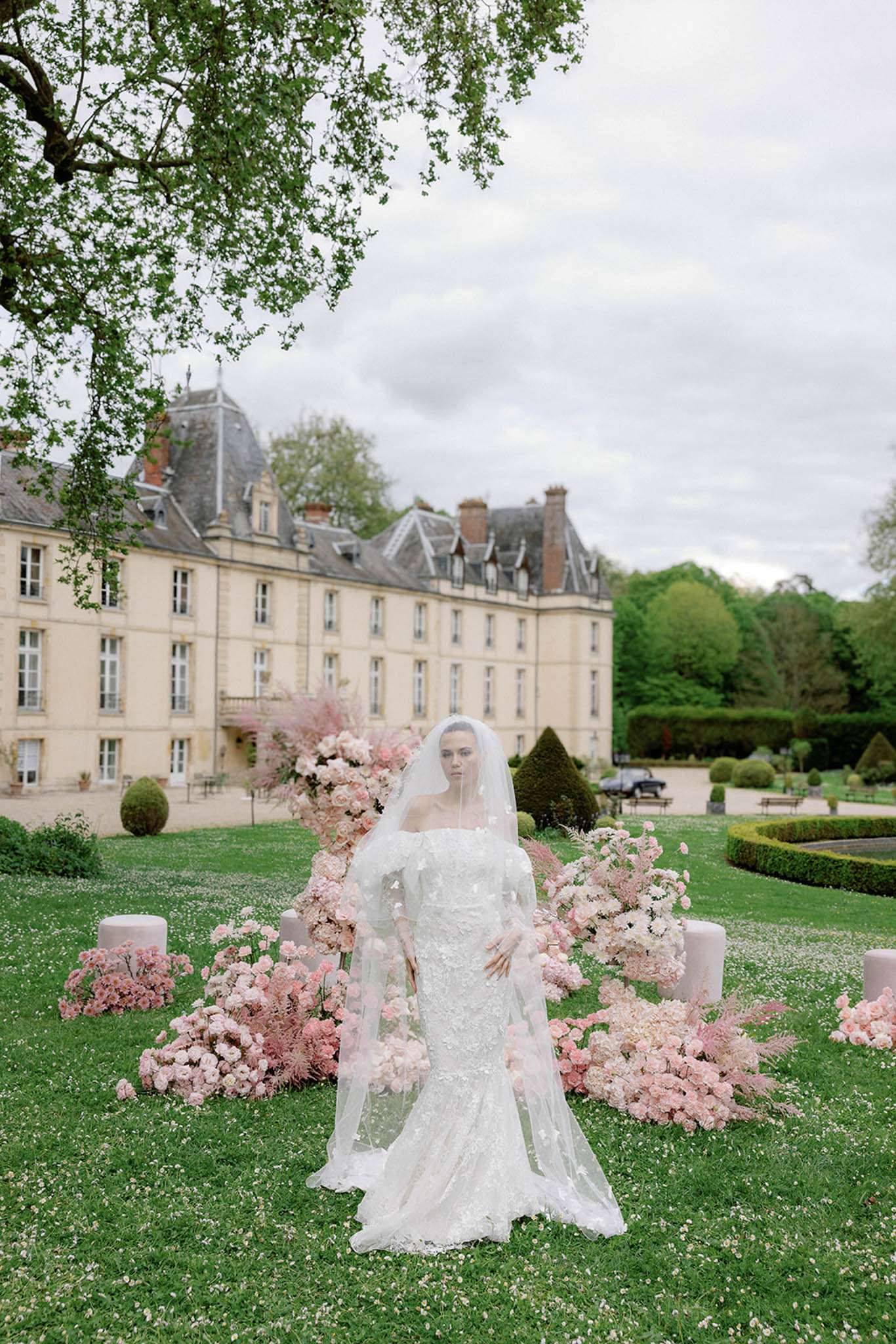 Bride in ivory lace gown with cathedral veil standing amid blush pink floral installation at chateau