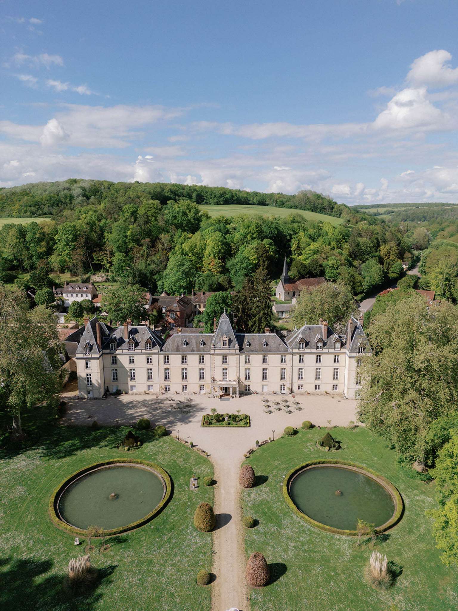 Aerial view of cream stone chateau with mansard roofs and twin circular reflecting pools on formal lawn