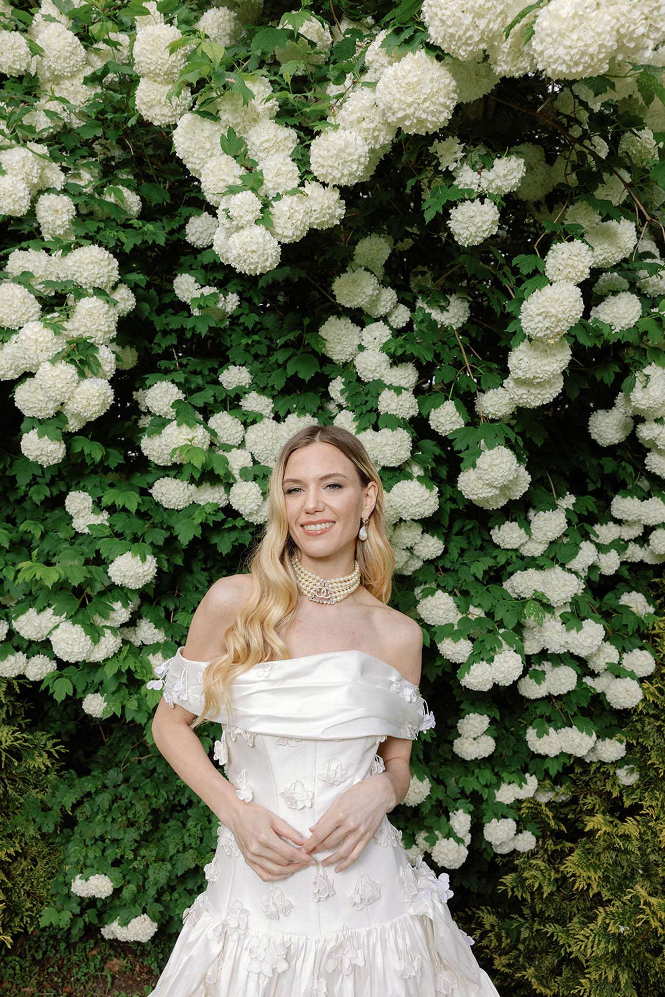 An outdoor bridal portrait of the bride standing alone in a garden setting. She wears an off-the-shoulder ivory gown with a draped bodice and three-dimensional floral appliqués scattered across the skirt, paired with a multi-strand pearl choker necklace with a decorative clasp at center front and drop pearl earrings. Her blonde hair falls in loose waves over her shoulders. The backdrop is a large flowering viburnum shrub covered in dense, globe-shaped white blooms against deep green foliage, which visually frames the bride. The composition is a mid-length portrait shot with the bride centered in the frame.