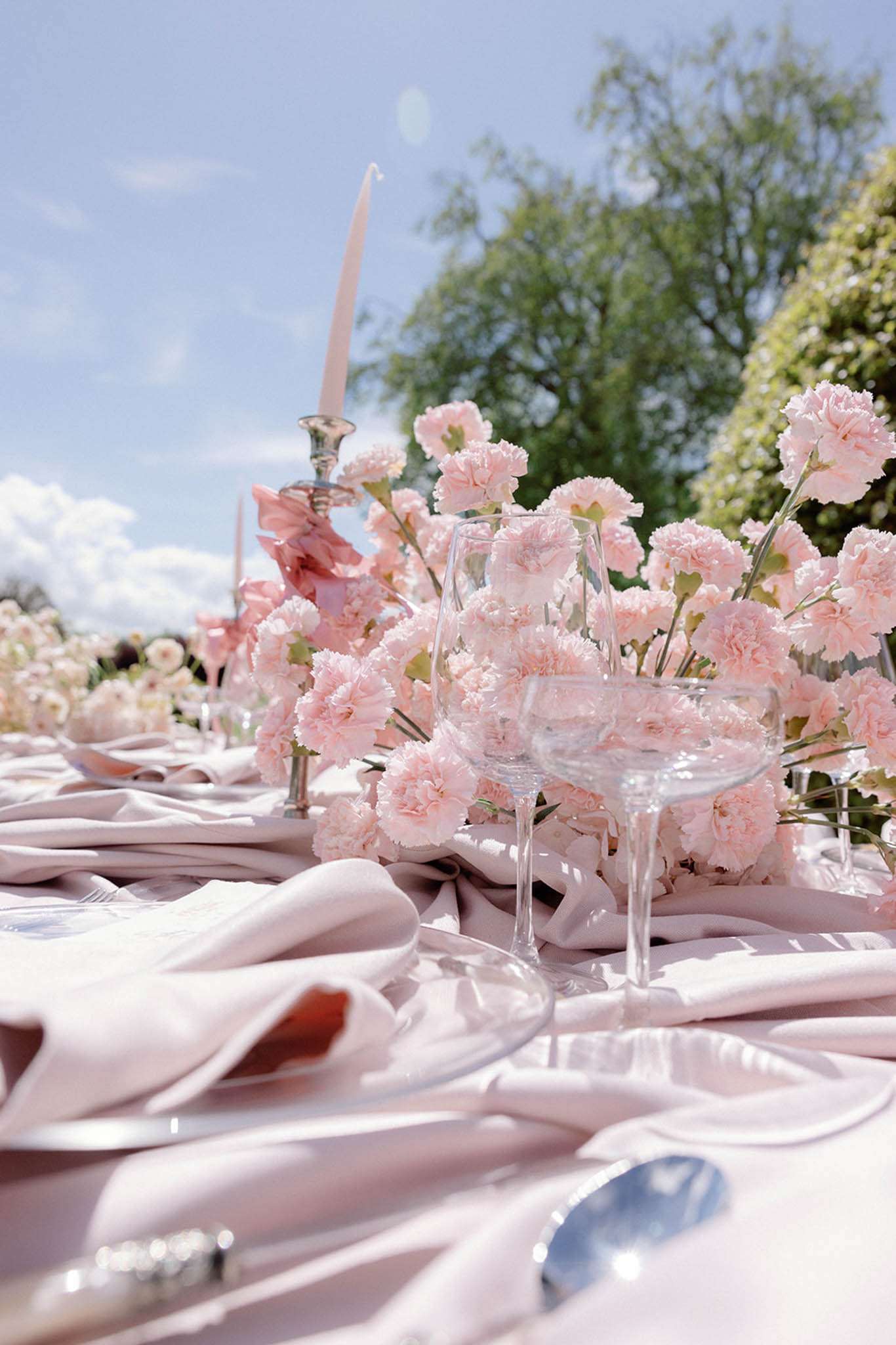 A close-up detail shot of an outdoor wedding reception table setting, photographed from a low angle at table level. The tablescape features blush pink carnation centerpieces arranged in full, dense clusters, paired with a tall blush taper candle in a silver candlestick holder. The table is dressed with draped blush pink linen napkins loosely gathered around the place settings, and clear crystal glassware including a wine glass and a coupe glass are visible in the foreground. Silver cutlery is partially visible at the edges of the frame. The overall decor palette is monochromatic blush pink, consistent with a romantic, soft, classic styling theme. The table extends into the background where additional place settings and floral arrangements are visible in soft focus.
