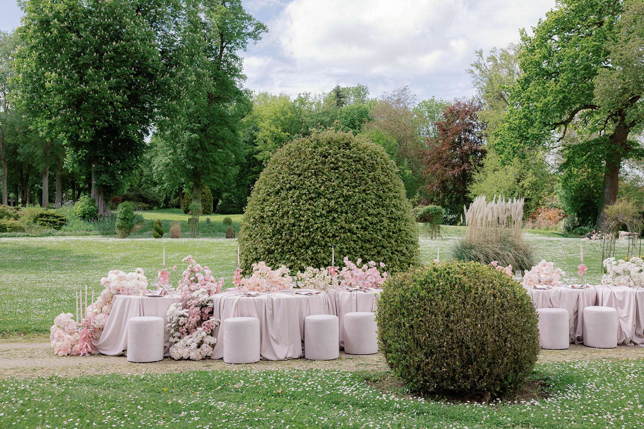 An outdoor wedding reception tablescape set in a formal garden with manicured topiary and mature trees. Two round tables are dressed in blush pink floor-length linen with cylindrical upholstered stools in the same blush tone used as seating. The centerpieces are abundant low-spread arrangements of blush pink ranunculus, white hydrangea, pale pink roses, and ivory blooms cascading down to ground level, with tall tapered pink candles interspersed throughout. The tables are positioned around a large dome-shaped topiary, integrating the formal garden landscaping as a design element. The overall color palette is a tonal blush and white scheme with a modern, romantic styling approach. Wide establishing shot capturing the full table setup within the garden setting.