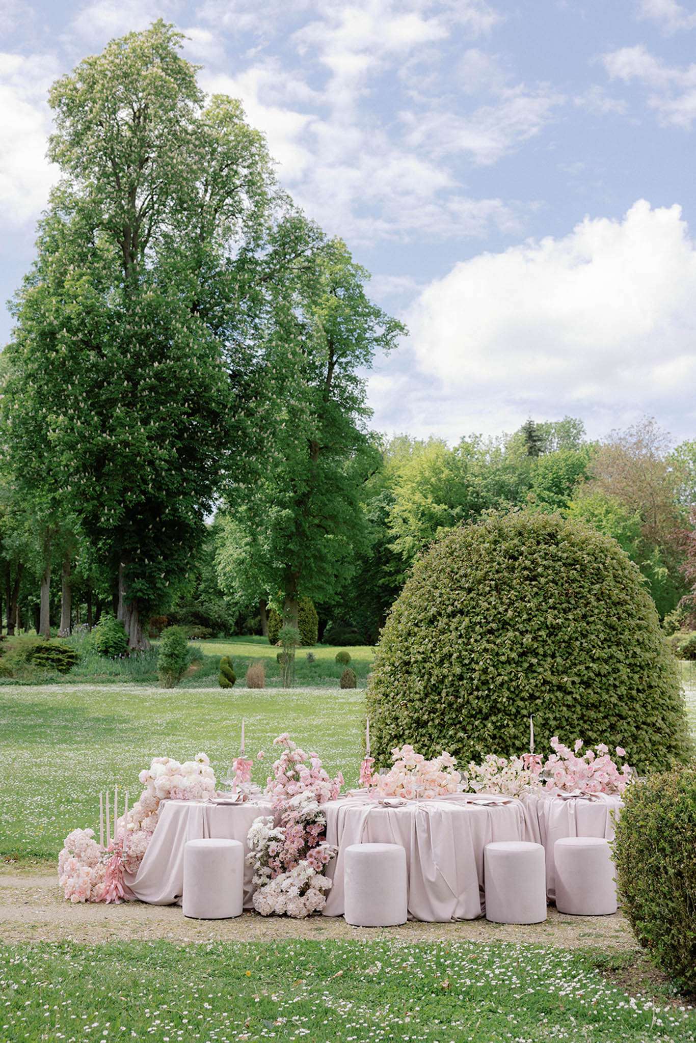 Round reception table in blush pink linen with overflowing pink roses and hydrangeas beside clipped topiary hedge