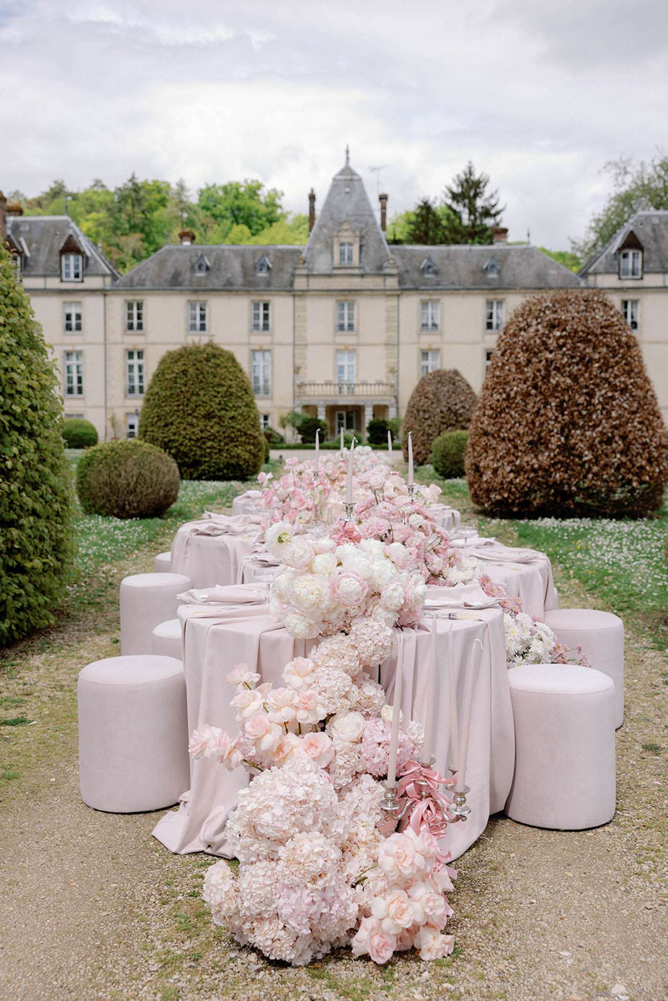 Long outdoor reception table with blush linen and overflowing pink and white floral runner in front of a French chateau