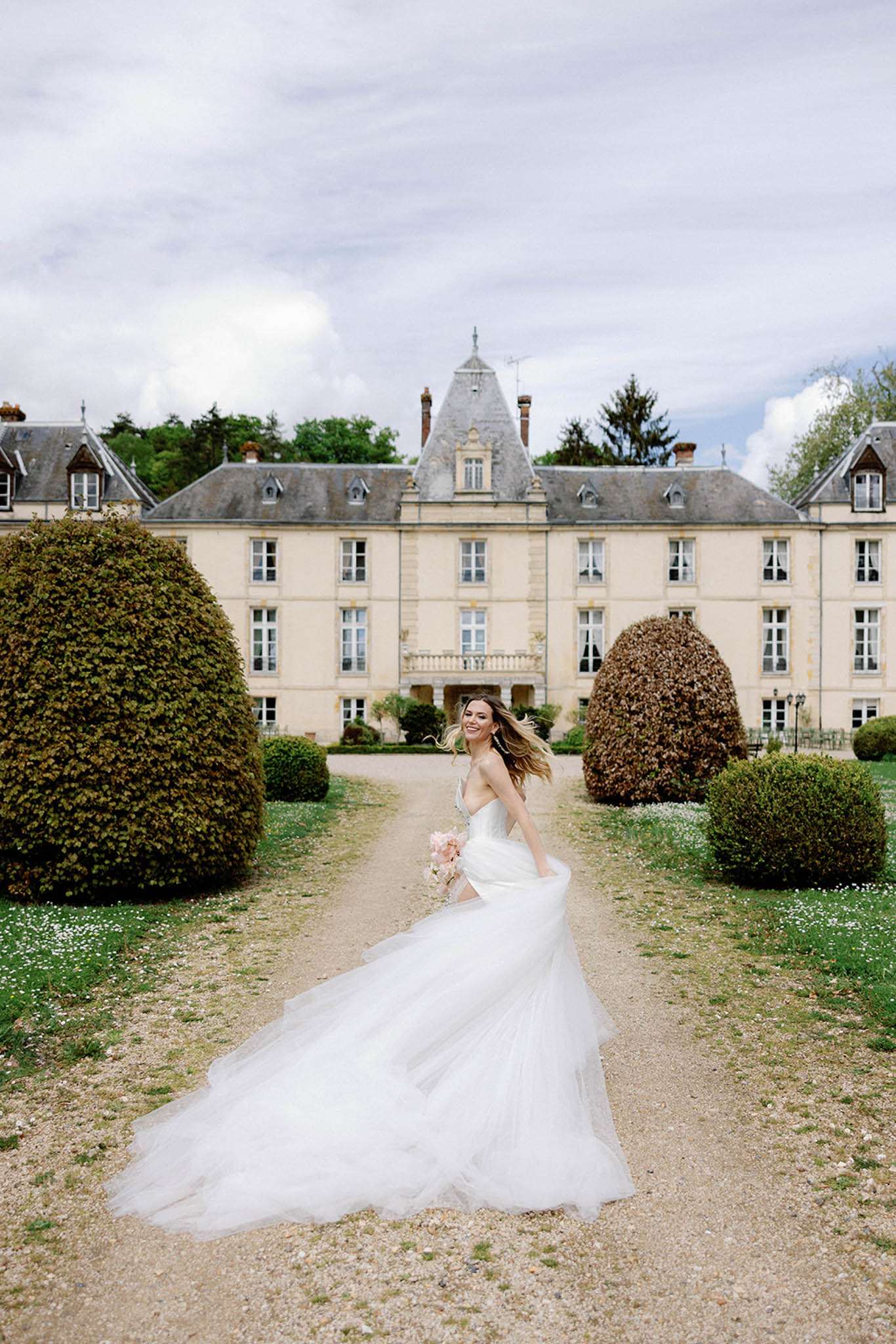 A bridal portrait taken outdoors on the gravel pathway leading up to a large French château, with the full facade of the multi-story cream-stone building visible in the background. The bride is mid-movement, turning toward the camera with her hair tossing in the wind, holding a small blush pink bouquet in one hand while gathering her voluminous white tulle ballgown skirt with the other. The dress features a fitted bodice with thin straps and a full layered tulle skirt with an extended train that fans out across the gravel path. Formally clipped topiary hedges frame either side of the pathway in the mid-ground. The shot is a full-length portrait taken from a low-to-mid angle, capturing both the bride and the full architectural backdrop of the château. Potential venue feature image.