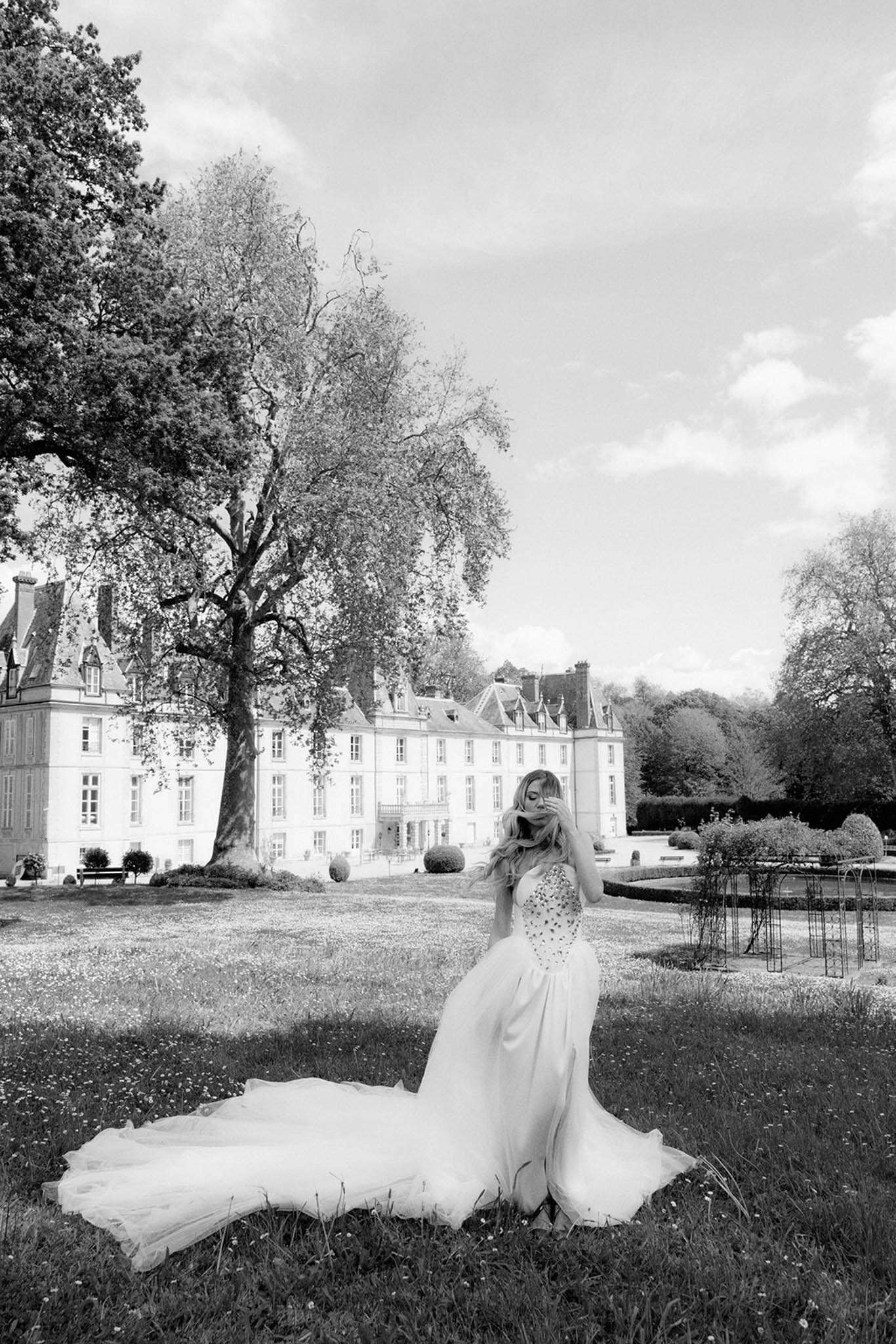 Black-and-white portrait of bride in strapless embellished ball gown seated on lawn before a French chateau