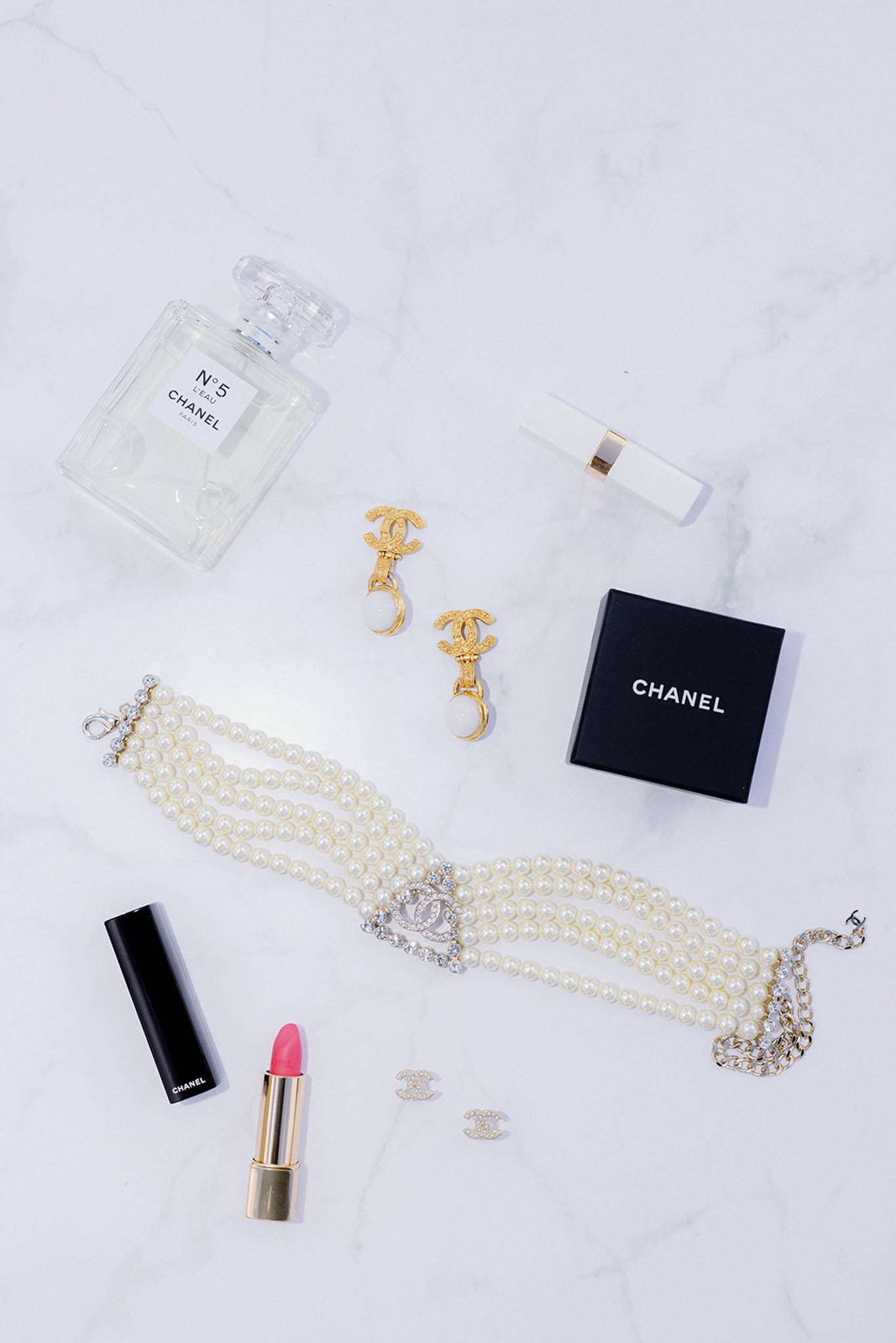 A flat lay detail shot of bridal accessories arranged on a white marble surface. Items include a Chanel Nº5 L'Eau perfume bottle, a white and gold Chanel lipstick case, a Chanel Rouge Allure lipstick in a coral-pink shade with a black and gold case, a small black Chanel branded box, a pair of gold Chanel CC drop earrings with pearl drops, a pair of small gold Chanel CC stud earrings, and a multi-strand pearl choker necklace featuring a crystal-encrusted CC logo centerpiece with a silver and crystal chain clasp. The styling is classic and polished, with a consistent gold, pearl, and white color palette throughout.