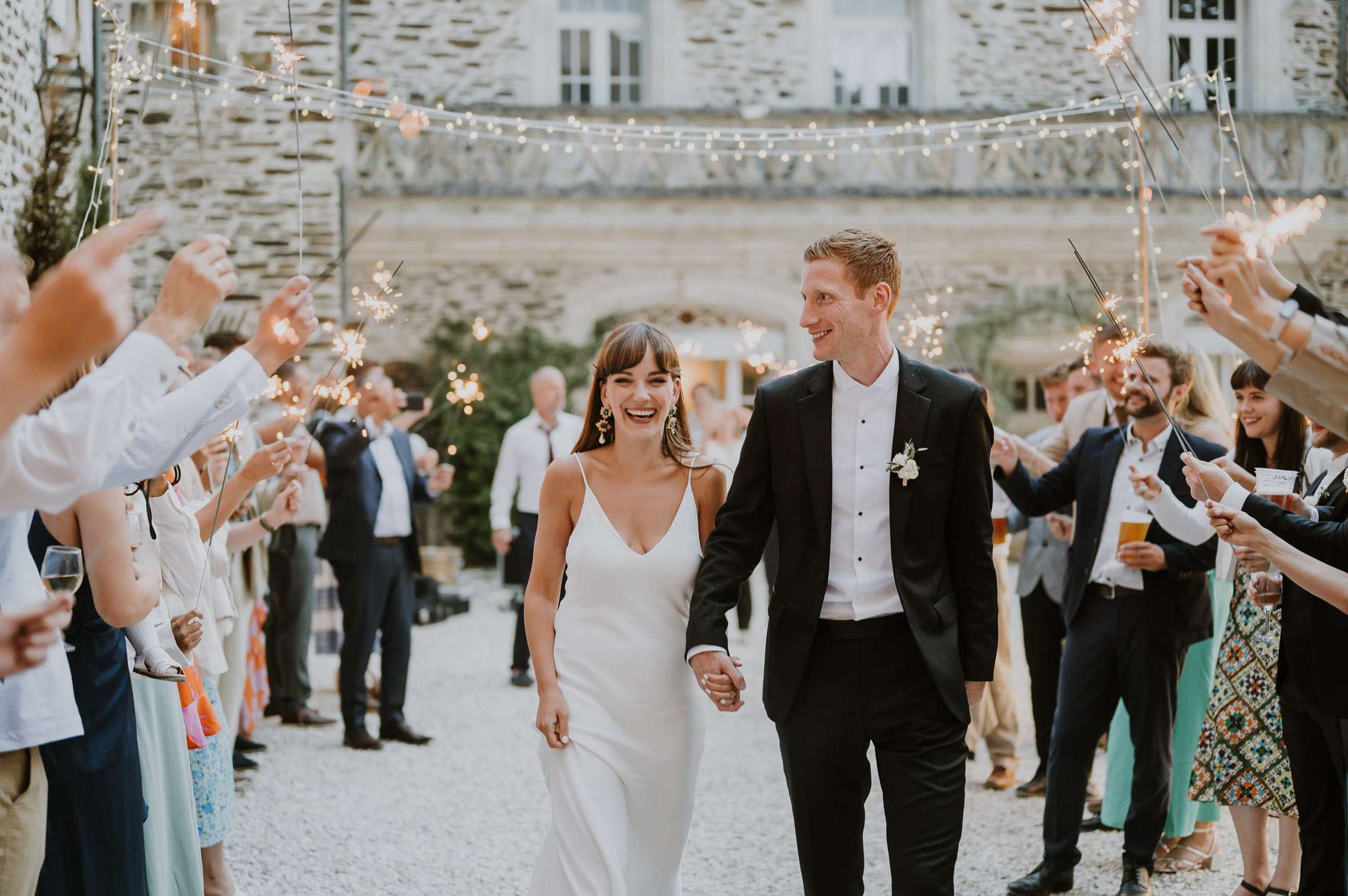 Bride and groom walk through a sparkler send-off tunnel in a chateau courtyard lit by fairy lights