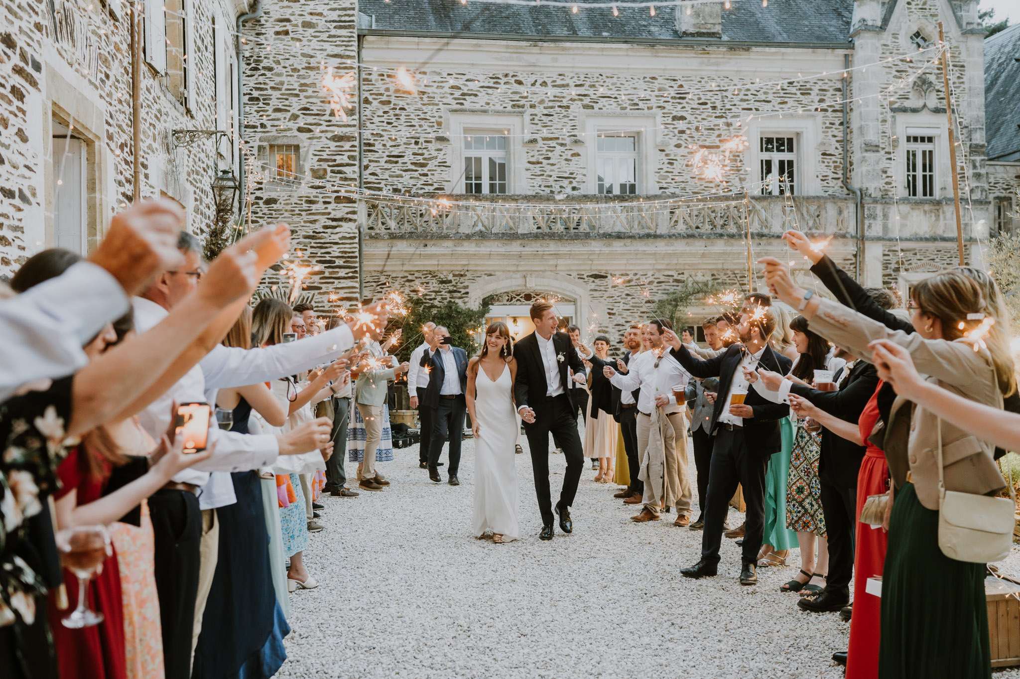 A sparkler send-off exit takes place in the gravel courtyard of a French château, with the couple walking hand-in-hand through a tunnel formed by approximately 40–50 guests holding lit sparklers on either side. The bride wears a simple white slip-style gown with a V-neckline, and the groom is dressed in a black suit with a white dress shirt, no tie. Fairy lights strung across the courtyard overhead add to the warm glow of the sparklers against the early evening light. The wide shot captures the full scene with the historic grey stone château façade as the backdrop, styled in a modern classic aesthetic.