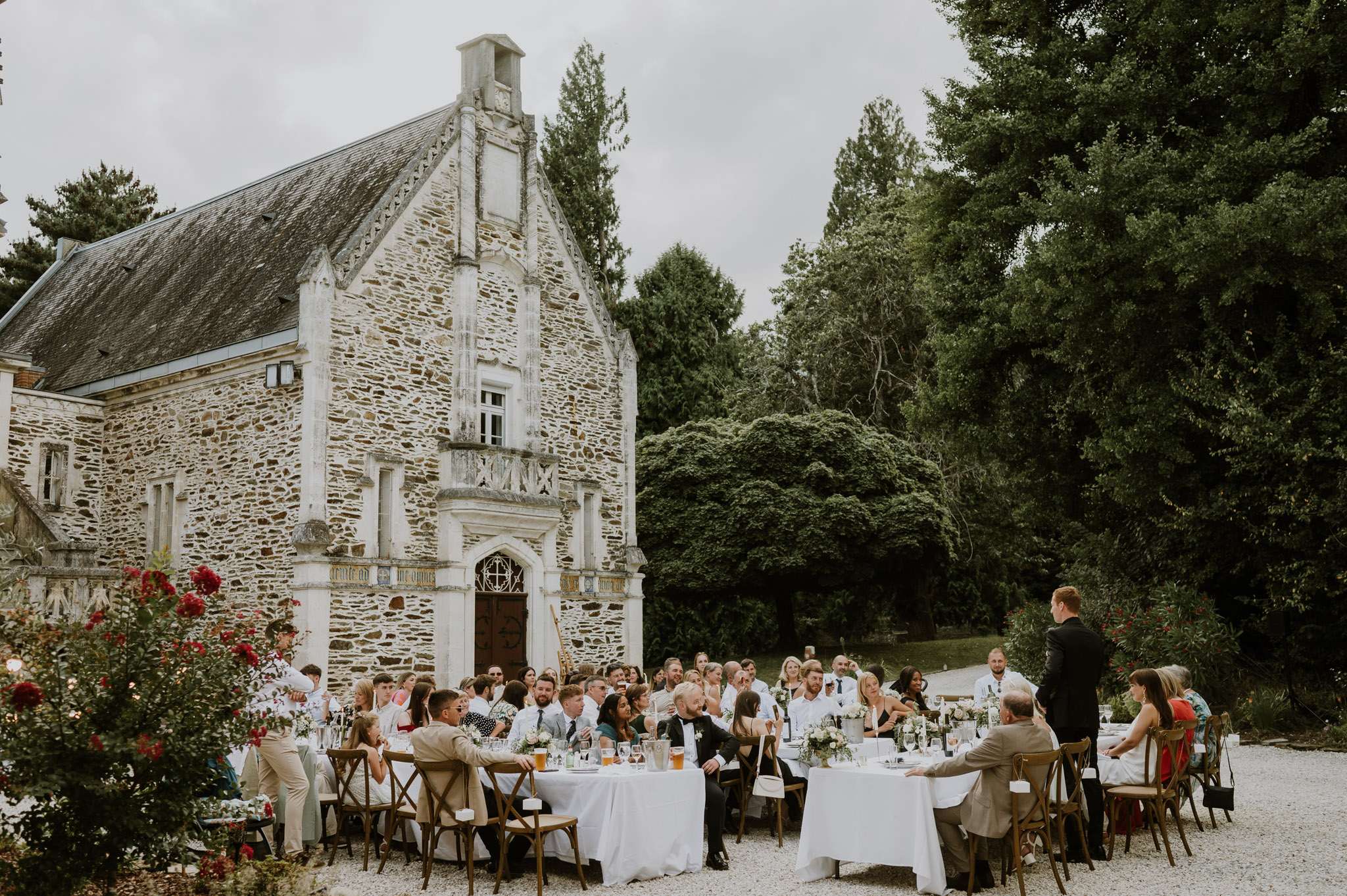 An outdoor wedding reception dinner is taking place on a gravel courtyard in front of a historic French stone manor or château with Gothic architectural details, including a pointed gabled facade, arched doorway, and mullioned windows. Approximately 50–60 guests are seated at round tables covered with white linen tablecloths, arranged with wooden cross-back chairs, and decorated with small white floral centerpieces, glassware, and bottles. A person standing at the far right appears to be delivering a speech or toast to the assembled guests. The overall decor style is classic and understated, with a white and green color palette, and the wide-angle shot captures both the full guest seating arrangement and the château facade as a backdrop. Potential venue feature image.