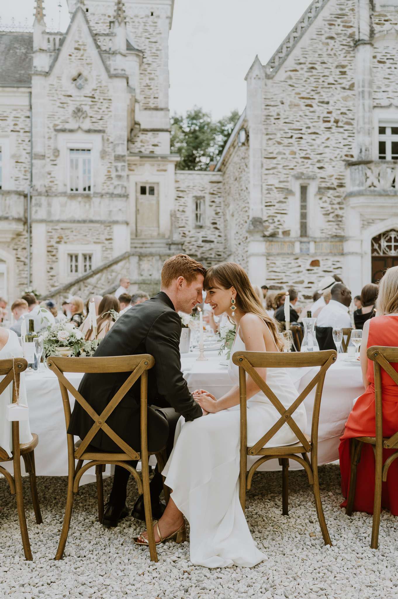 Couple leans foreheads together at candlelit reception table in gravel courtyard of Gothic-turret chateau