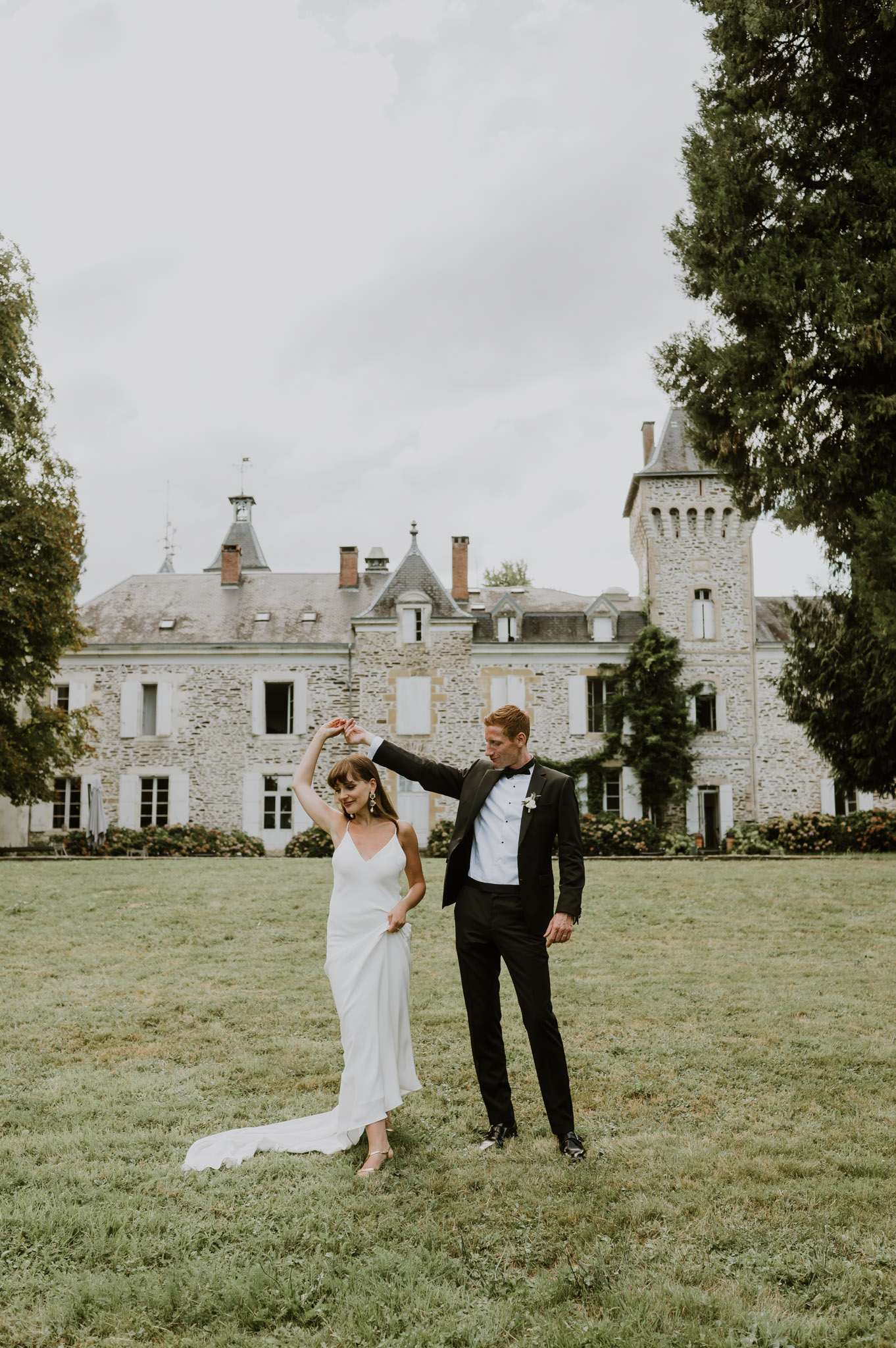 Groom spinning bride on chateau lawn with turret-topped manor in background, both in modern minimal wedding attire
