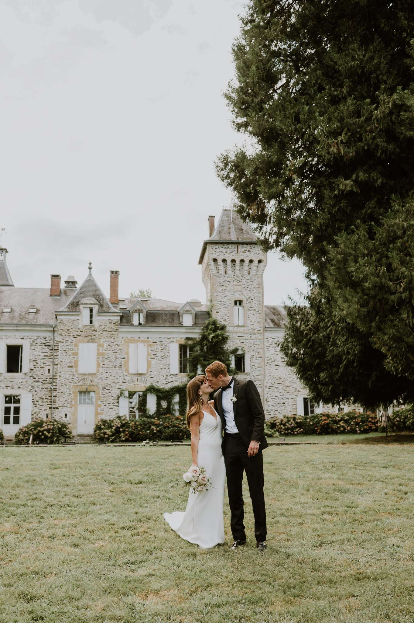 Bride and groom kissing on lawn in front of stone chateau with crenellated tower and climbing vines