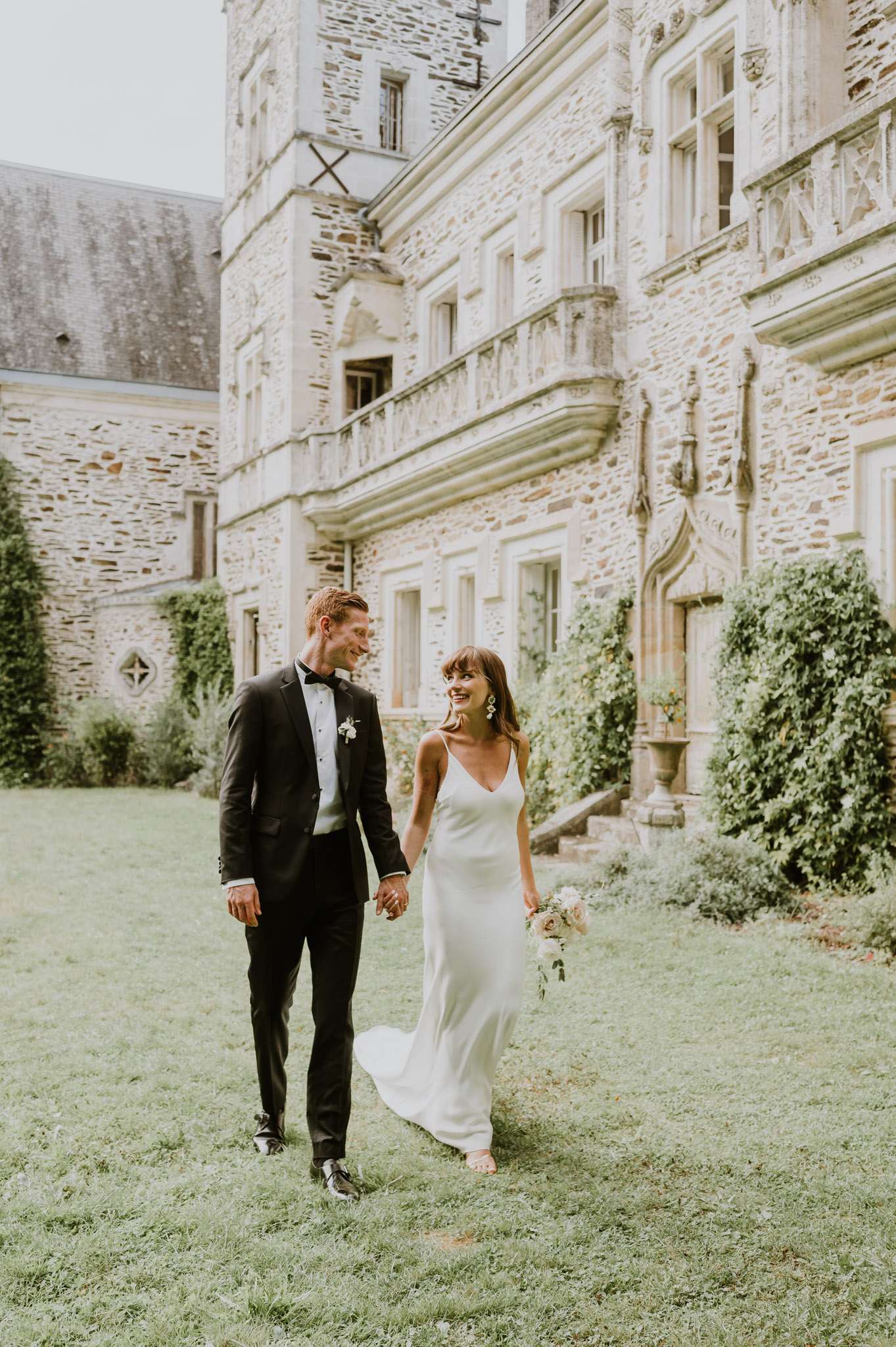 A couple portrait taken outdoors on the grounds of a French château, with the couple walking hand-in-hand across a lawn in front of the multi-story stone facade. The groom wears a black tuxedo with a black bow tie and a small white floral boutonniere, while the bride wears an ivory silk slip dress with thin spaghetti straps and a V-neckline, accessorized with drop earrings. She carries a loose bouquet of blush and cream flowers with greenery. The styling is modern and minimal against the historic Renaissance-style château architecture. The shot is a full-length portrait with a slightly warm, airy edit. Potential venue feature image.