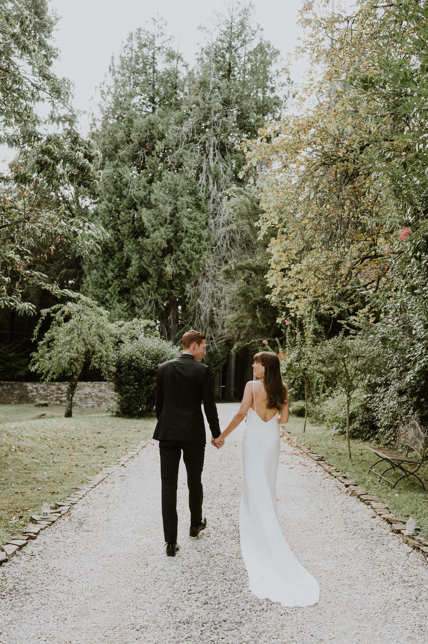 A couple's portrait taken outdoors on a gravel pathway lined with mature trees and sculpted shrubs, likely within the grounds of a French estate or château. The couple is walking away from the camera hand-in-hand, with the bride glancing back over her shoulder toward the groom. The bride wears a slim-fit ivory slip-style gown with thin spaghetti straps, an open low back, and a short train; the groom wears a fitted black suit. The styling is modern and minimal with no visible bouquet or accessories. The shot is a full-length wide portrait framed centrally along the path, with a dark ivy-covered structure visible in the background.