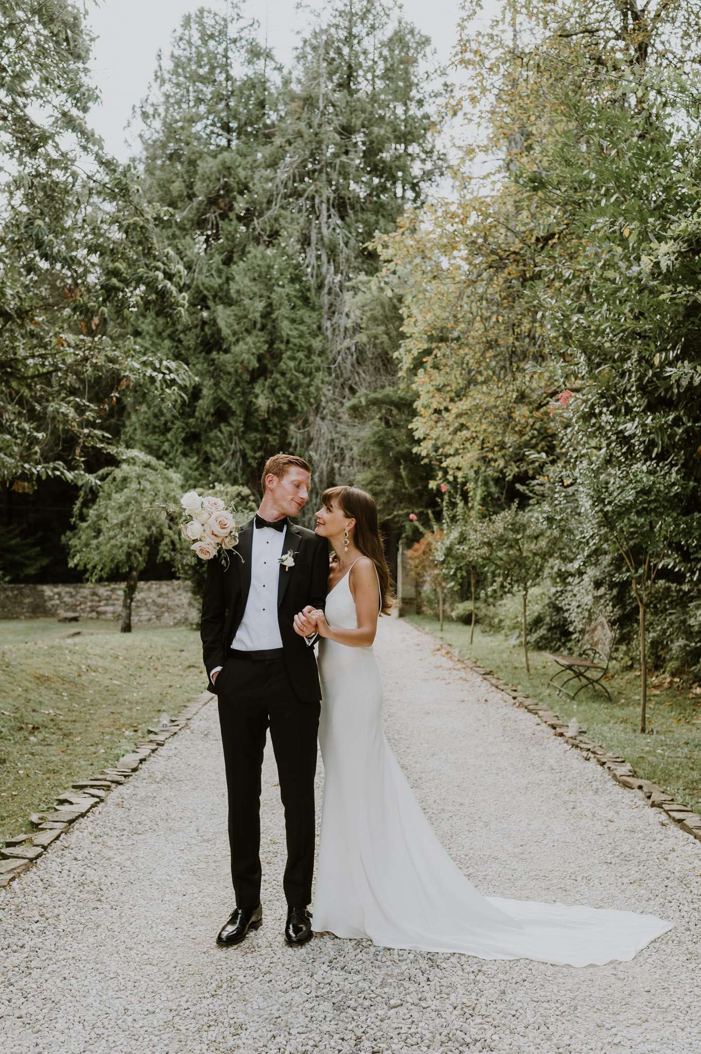 A couple portrait taken outdoors on a gravel path edged with stone borders, set within a wooded garden. The groom wears a black tuxedo with a black bow tie and a small white floral boutonniere, while the bride wears a white spaghetti-strap slip-style gown with a long train trailing behind her. The bride carries a bouquet of blush and cream roses and leans into the groom, with both looking at each other and smiling. The overall styling is classic and minimal, with a neutral color palette of black, white, and blush. This is a full-length couple portrait shot.