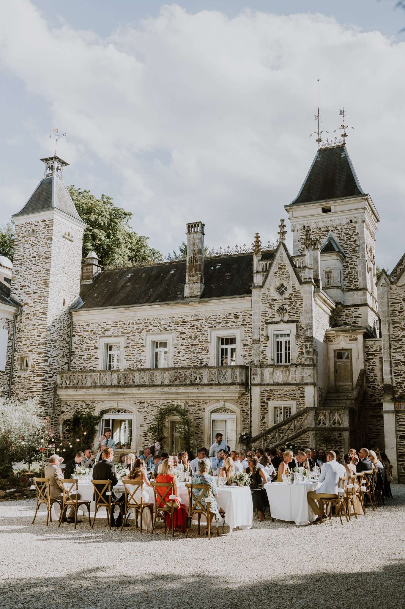 An outdoor wedding reception dinner is taking place in the courtyard of a French château, with approximately 50–60 guests seated at round and long rectangular tables covered in white linen. The tables are decorated with low floral centerpieces featuring white blooms and greenery, and string lights are visible above the seating area. Guests are dressed in a mix of formal and semi-formal attire, with notable pops of color including a red dress and a coral/pink dress among the seated guests. The seating uses natural wood cross-back chairs throughout, consistent with a relaxed classic style. The château itself is a multi-story French stone manor with two flanking towers topped with dark pointed roofs, ornate Gothic-style stonework, arched ground-floor doorways, and an exterior stone staircase. Wide shot capturing both the full architectural facade and the entire reception layout. Potential venue feature image.