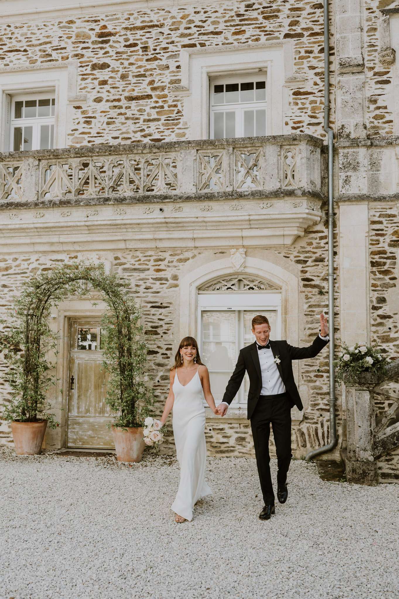 Bride with blush bouquet and groom in black tuxedo walking across chateau courtyard with carved balustrade