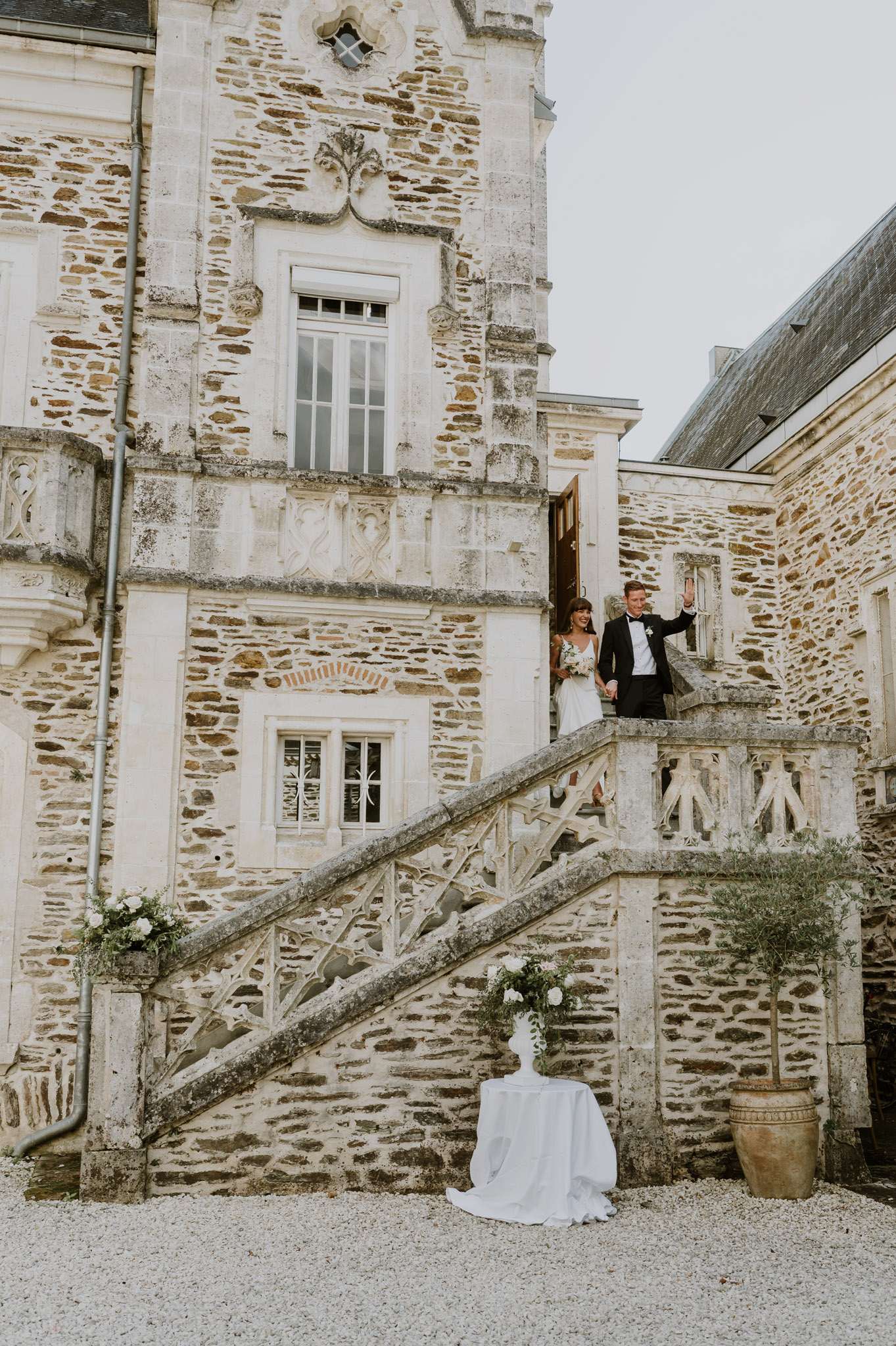 Bride and groom emerge from a chateau doorway atop an ornate stone staircase decorated with white floral arrangements