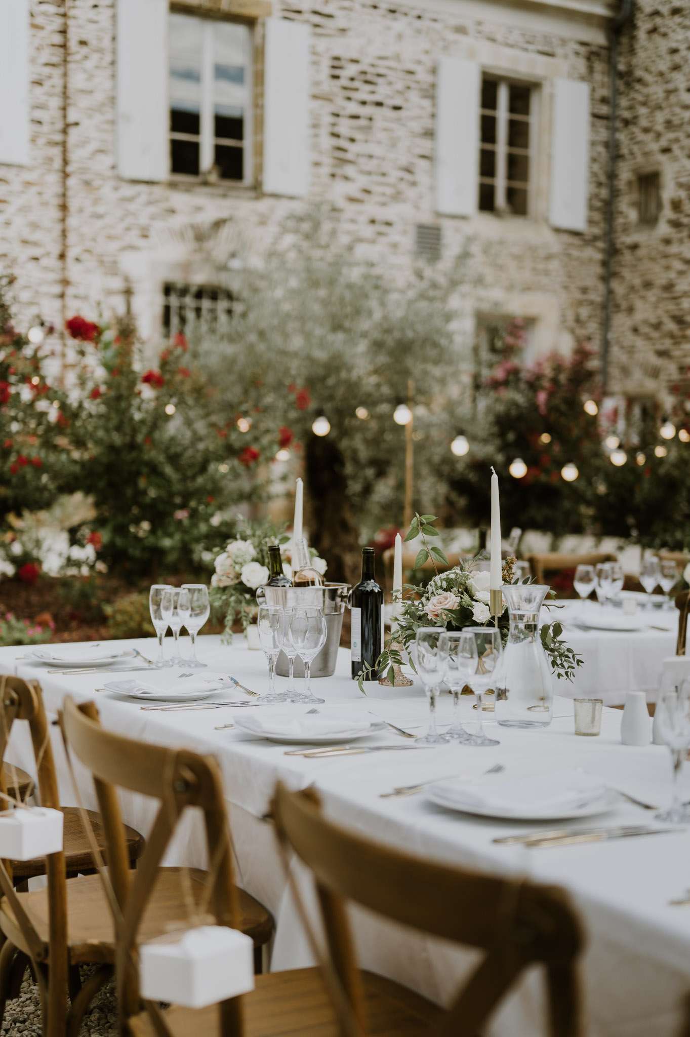 An outdoor wedding reception table setting photographed in medium close-up, positioned in a courtyard in front of a French stone manor house with white shuttered windows. The long rectangular table is dressed in a white linen tablecloth and set with white ceramic plates, silver cutlery, and crystal wine glasses, with a silver champagne bucket holding a bottle of white wine alongside an open bottle of red wine. Centerpieces consist of low arrangements of blush pink roses and white blooms with trailing greenery, flanked by tall ivory taper candles in gold holders. Natural wood cross-back chairs surround the table, and small white gift boxes are placed at each seat. String globe lights are strung between trees in the background, adding warm ambient lighting to the classic French garden setting with a romantic, understated décor palette of white, blush, and gold.
