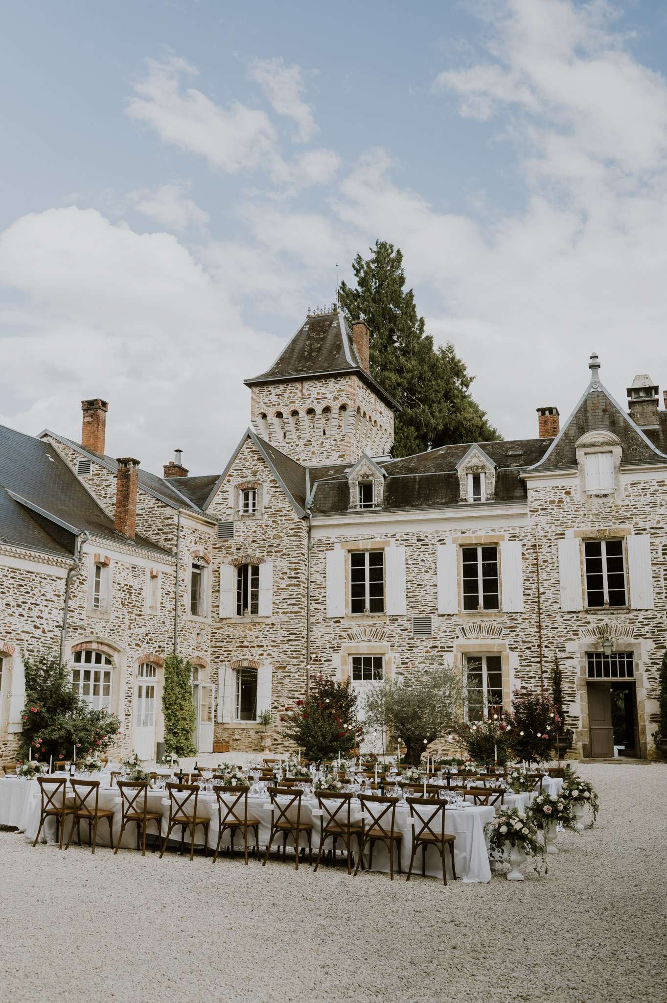 Outdoor reception with long banquet tables, cross-back chairs, and floral arrangements in a stone chateau courtyard