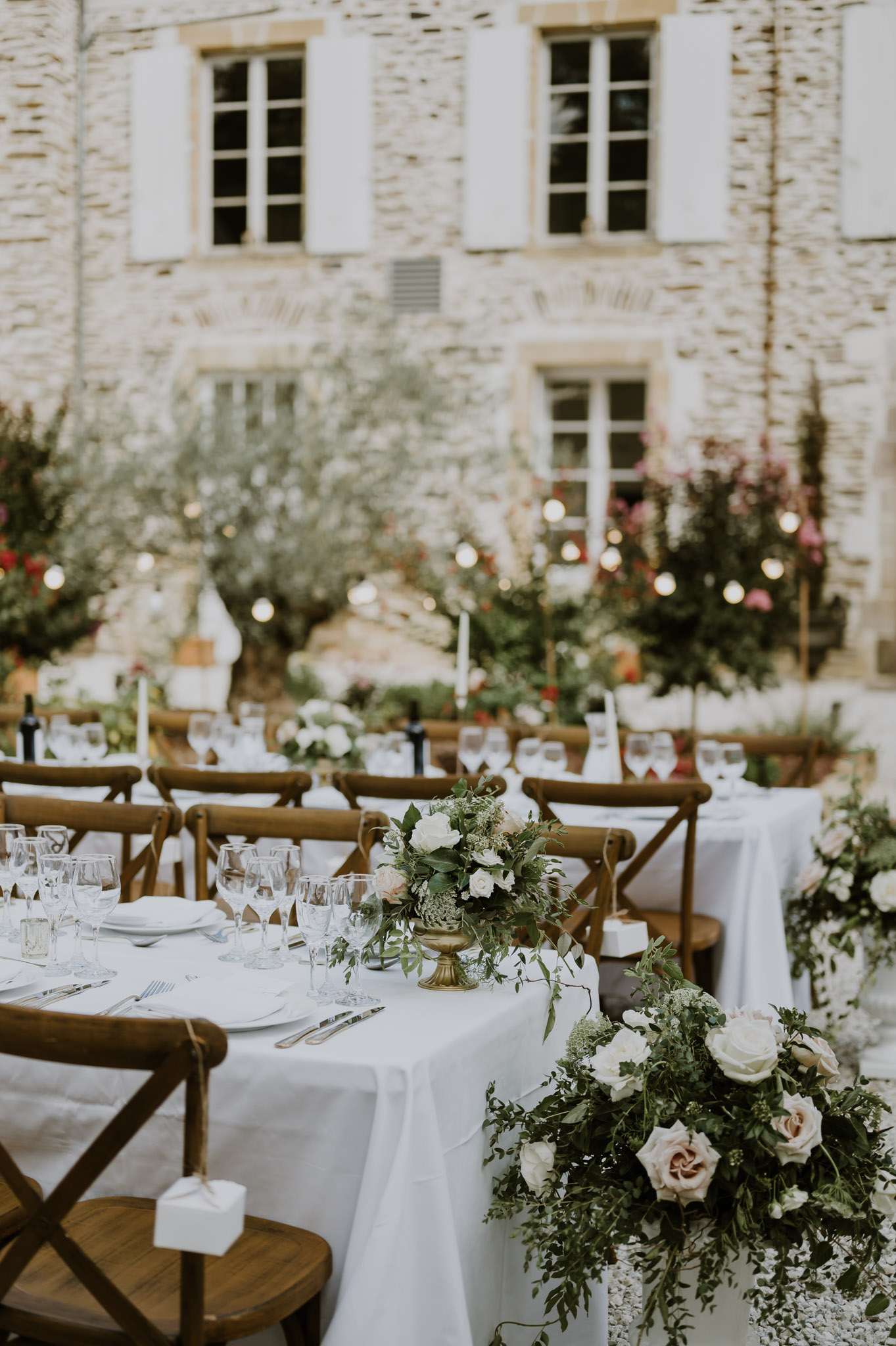 Long reception table with white linen, brass floral urns with trailing greenery and roses, and string lights