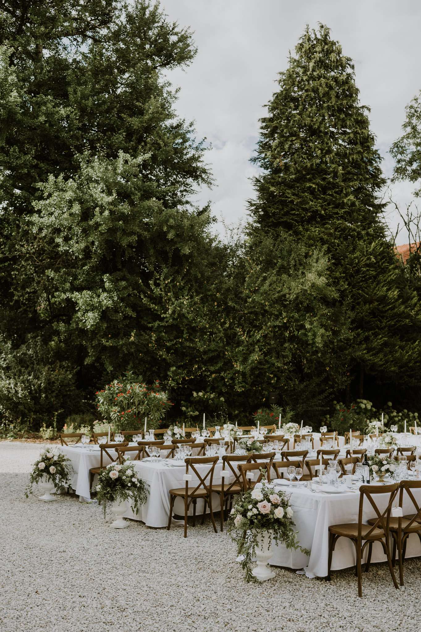 L-shaped long tables with blush rose arrangements in white urns and crossback chairs on white gravel
