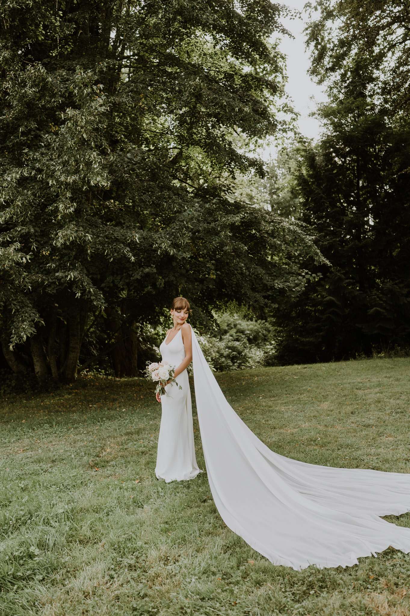 A bridal portrait taken outdoors on a lawn surrounded by mature trees. The bride stands alone, looking downward with a slight smile, wearing a minimalist ivory slip-style gown with thin straps and a very long cathedral-length veil that extends across the grass in a wide arc to the right of the frame. She holds a loosely arranged bouquet of blush pink and white flowers, likely peonies and roses, with soft greenery. The overall styling is clean and modern with a relaxed feel. The shot is a full-length portrait with the sweeping veil providing strong visual movement across the composition.