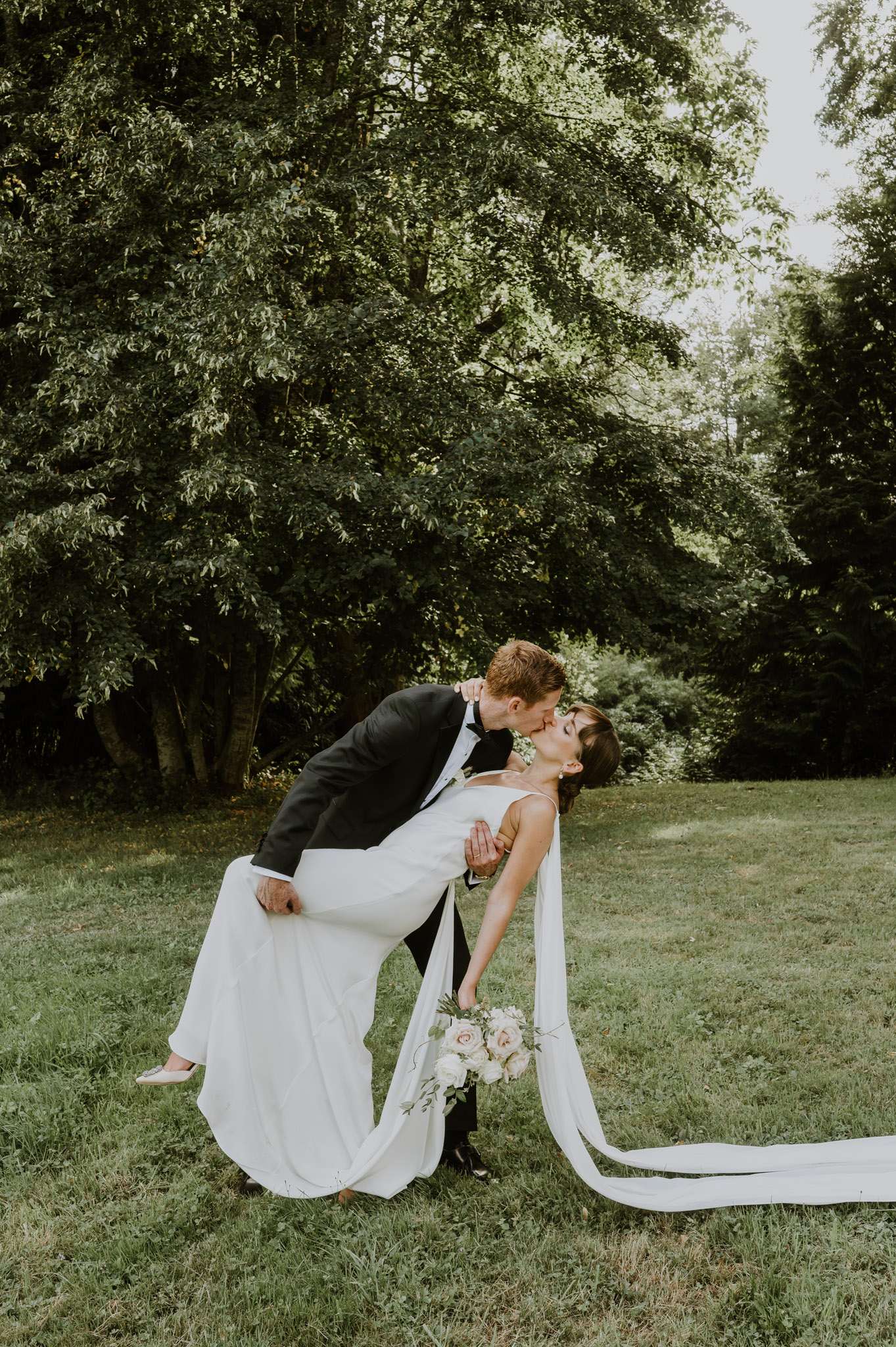 Groom dipping bride for a kiss on garden lawn, bride holding blush rose bouquet with trailing ribbon