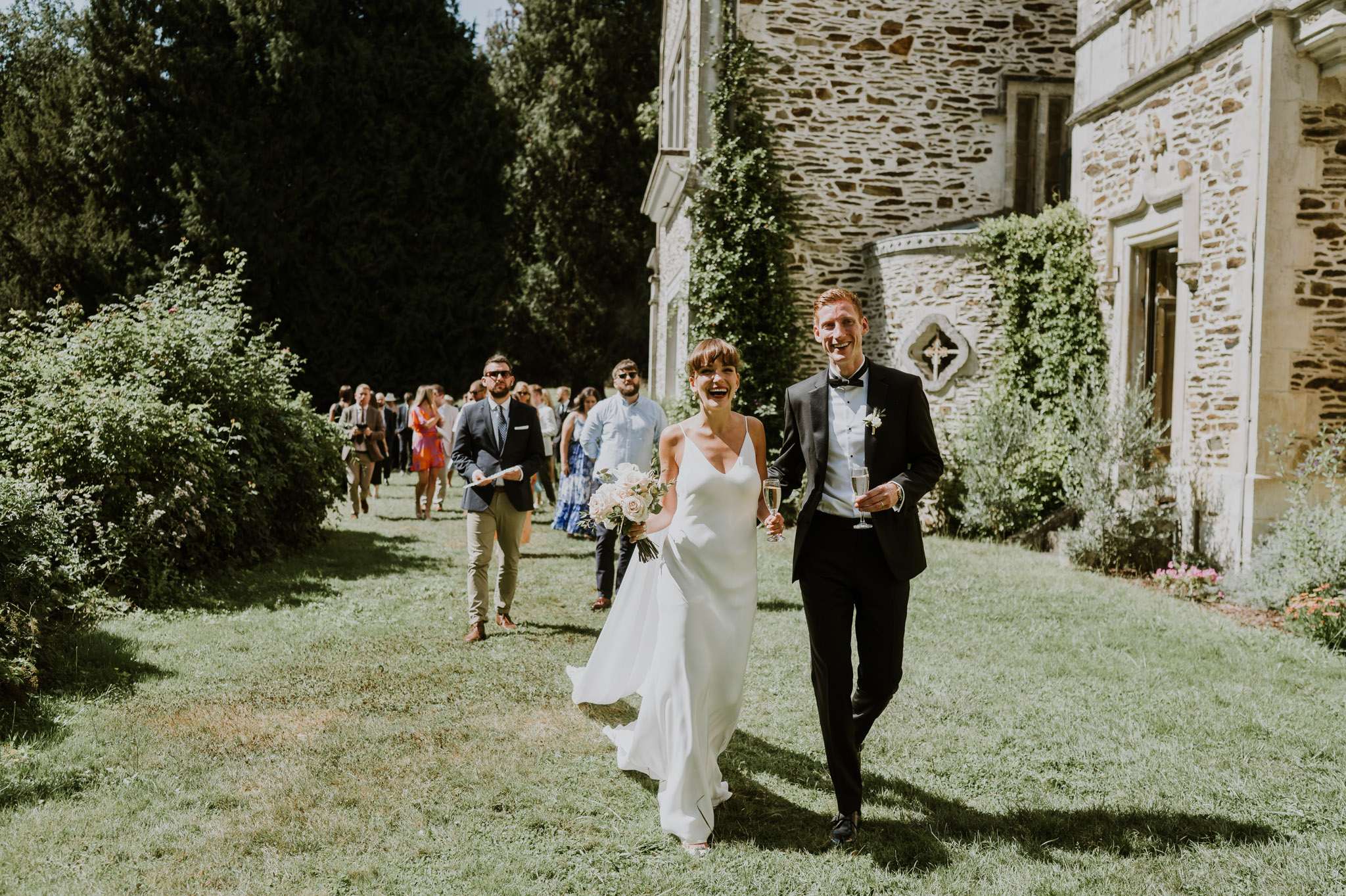 Couple leading 20 guests across lawn beside ivy-covered chateau, groom with champagne flute