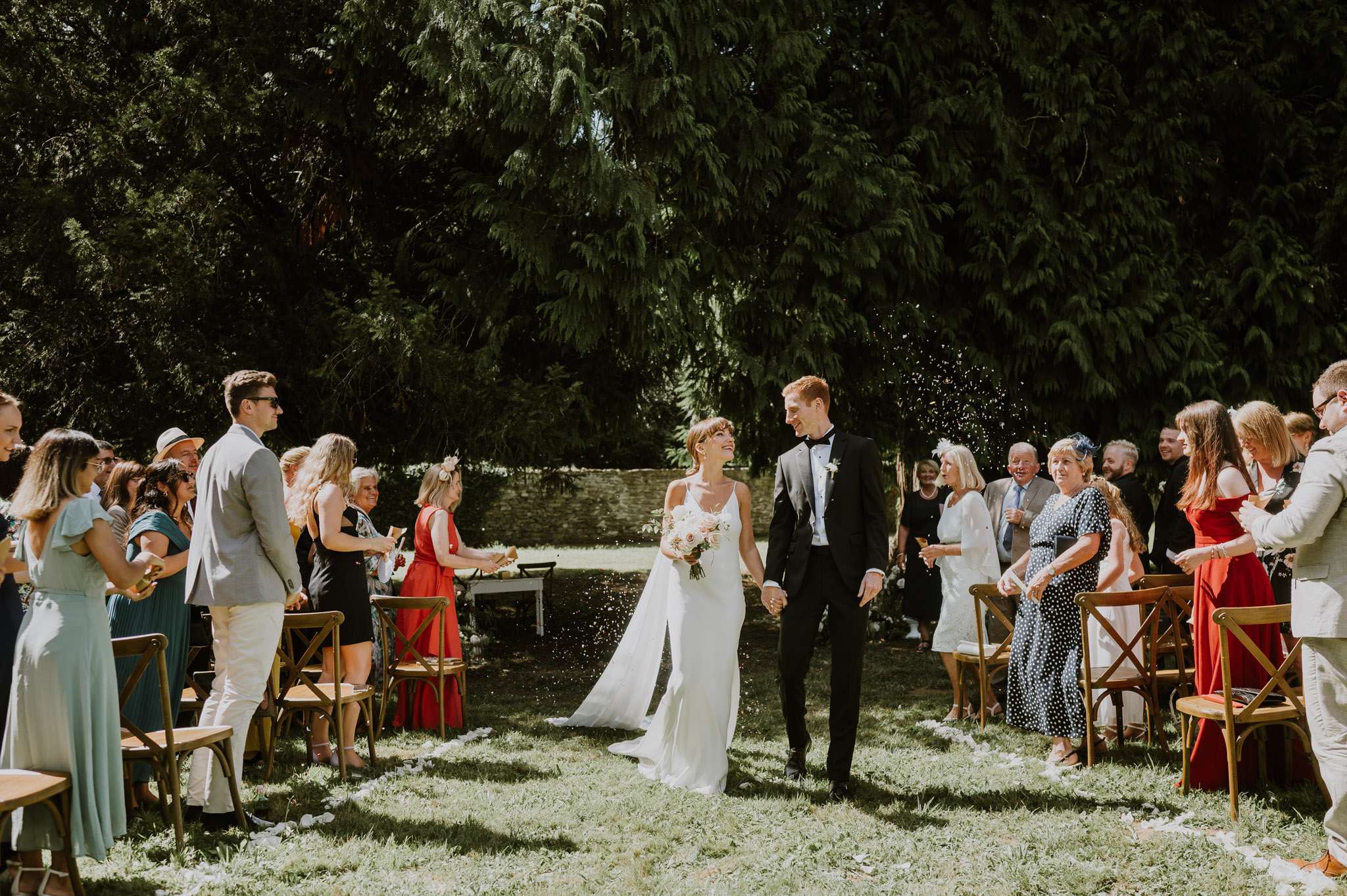 The bride and groom walk back down the aisle together immediately after their outdoor ceremony, holding hands and smiling at each other as guests on both sides throw white confetti or rice. The ceremony takes place in a garden setting with wooden cross-back chairs arranged in rows on a grass lawn, bordered by a low stone wall in the background. The bride wears a slim-cut white spaghetti-strap gown with a short train and carries a bouquet of blush pink and ivory flowers, while the groom wears a classic black tuxedo with a white boutonnière. Approximately 30–40 guests are visible, dressed in a mix of colorful summer outfits including red, teal, and navy, and the overall styling is classic with relaxed garden-party touches. The image is a wide mid-shot taken at ground level, capturing the full aisle scene with strong natural sunlight and deep tree shadows in the background.