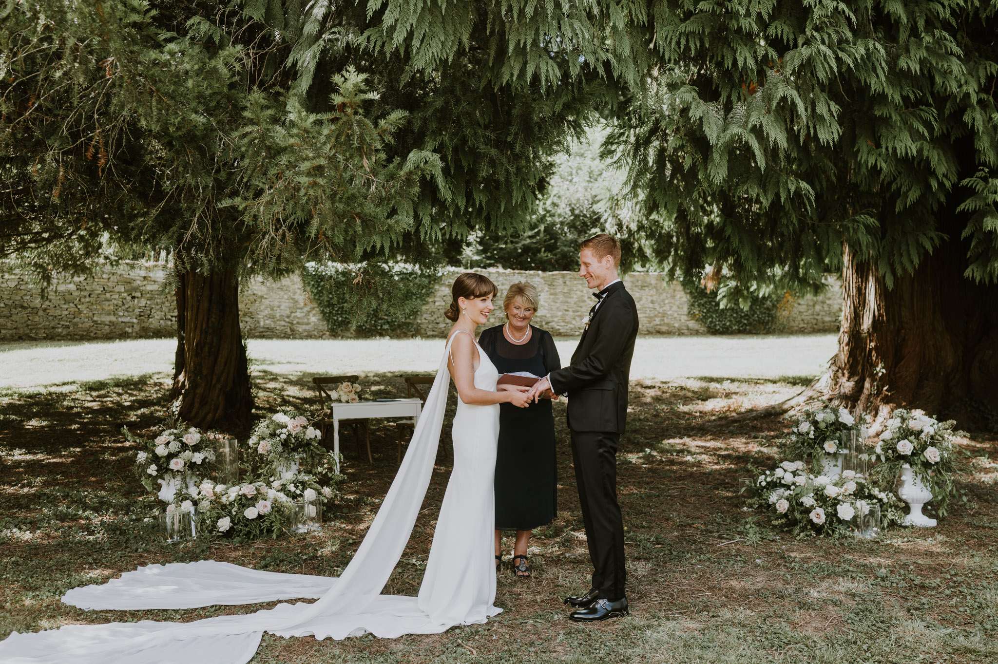 Bride in cape veil and groom in tuxedo holding hands during outdoor ceremony under mature trees in walled garden