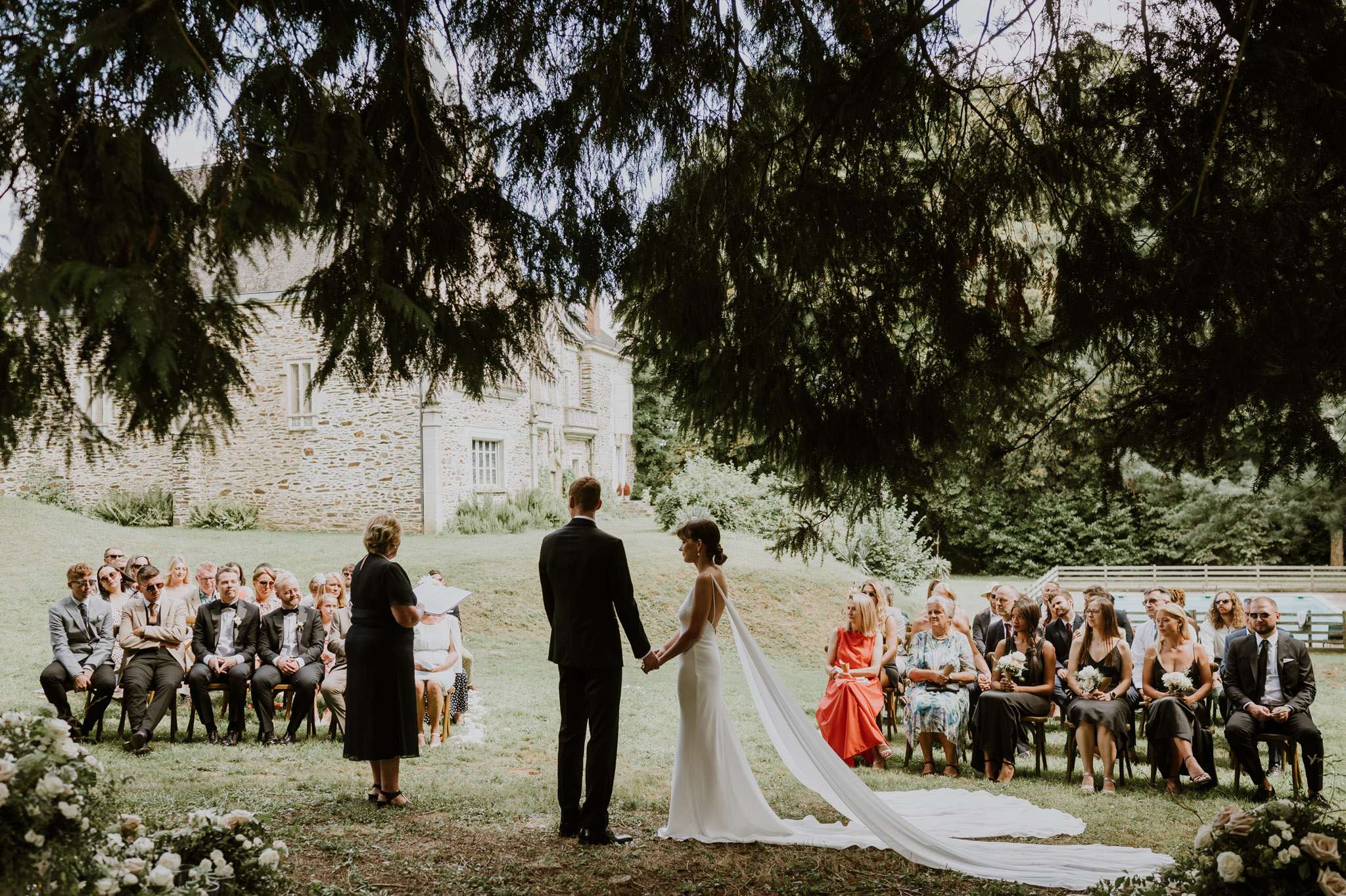 An outdoor wedding ceremony taking place on the grounds of a stone manor house or small château, with approximately 40–50 guests seated in wooden chairs arranged in two sections on either side of an aisle. The couple stands at the front holding hands, facing an officiant dressed in black who is reading from papers. The groom wears a dark navy suit, and the bride wears a fitted white sleeveless gown with a long cathedral-length veil trailing across the lawn. White and cream floral arrangements, including roses and greenery, are visible in the foreground at the base of the ceremony space. Guests are dressed in a mix of formal and semi-formal attire, with notable pops of coral and black among the crowd. The ceremony setup has a relaxed, classic outdoor style with no visible arch or structured altar. The shot is taken from a wide-angle perspective from behind the couple, framed by overhanging tree branches, with the stone manor building visible in the background.
