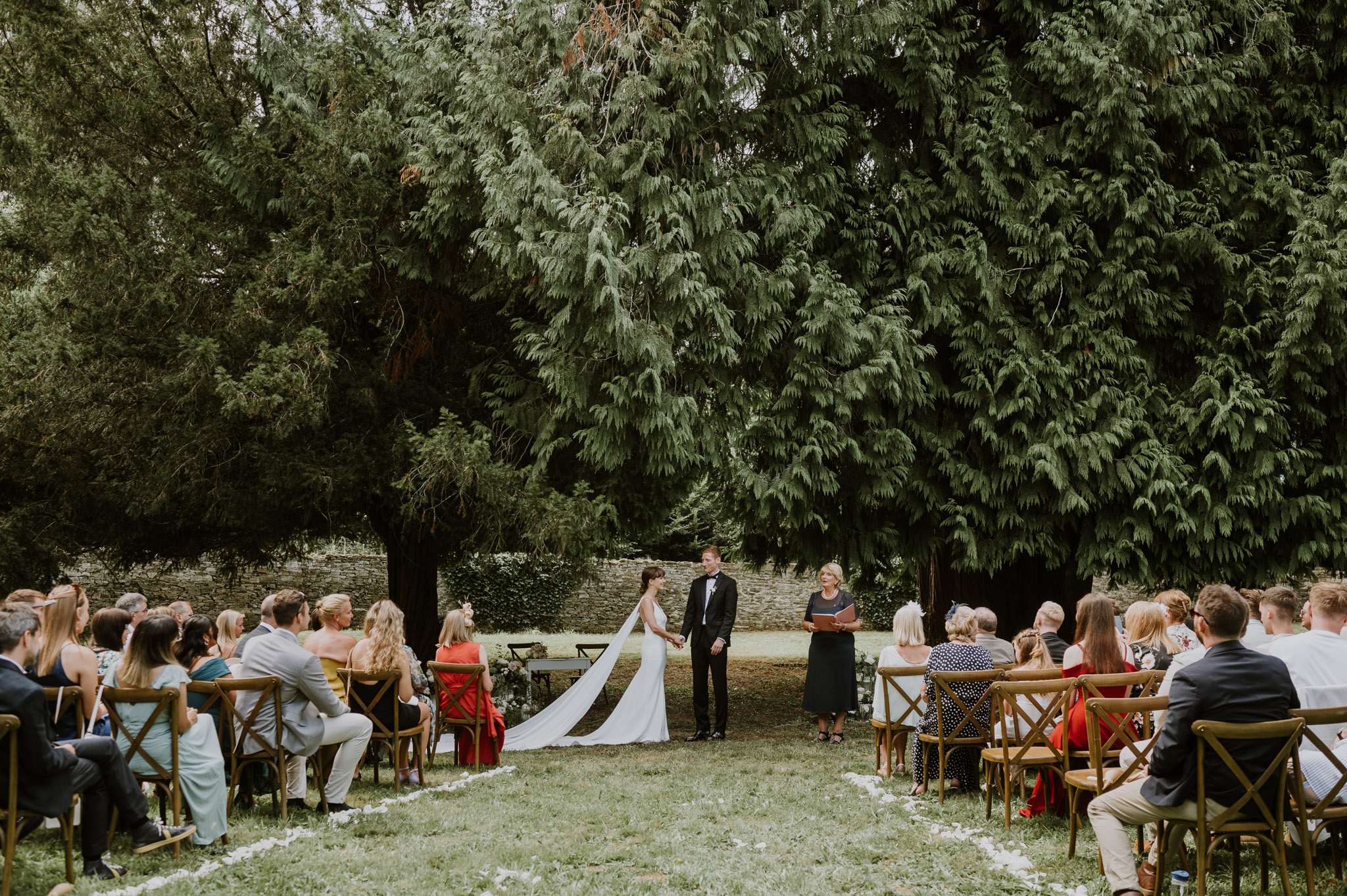 An outdoor wedding ceremony taking place on a lawn, with the couple standing at the altar facing a female officiant who holds a red book. The bride wears a slim white gown with a long cathedral-length veil, and the groom is dressed in a black tuxedo with a bow tie. Approximately 40–50 guests are seated in wooden cross-back chairs arranged in a curved semicircle, with a petal-lined aisle leading to the altar. Guest attire is colorful and includes red, teal, mustard, and mint outfits. The setting is an outdoor garden with a low stone wall visible in the background. The ceremony area has a relaxed, natural aesthetic with no visible floral arch or altar structure. The shot is a wide-angle view capturing the full ceremony layout from behind the seated guests.