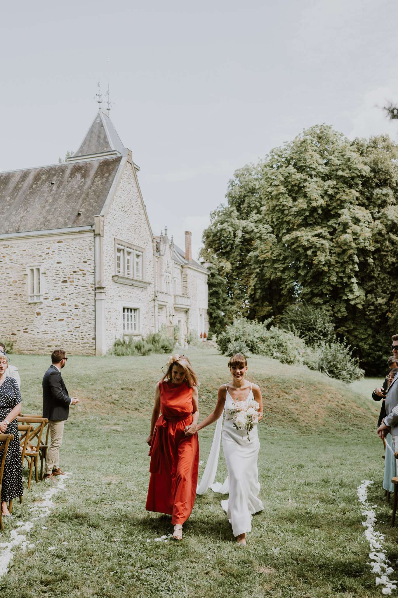 Bride in ivory slip gown walks petal-lined aisle with companion in red dress before stone chateau