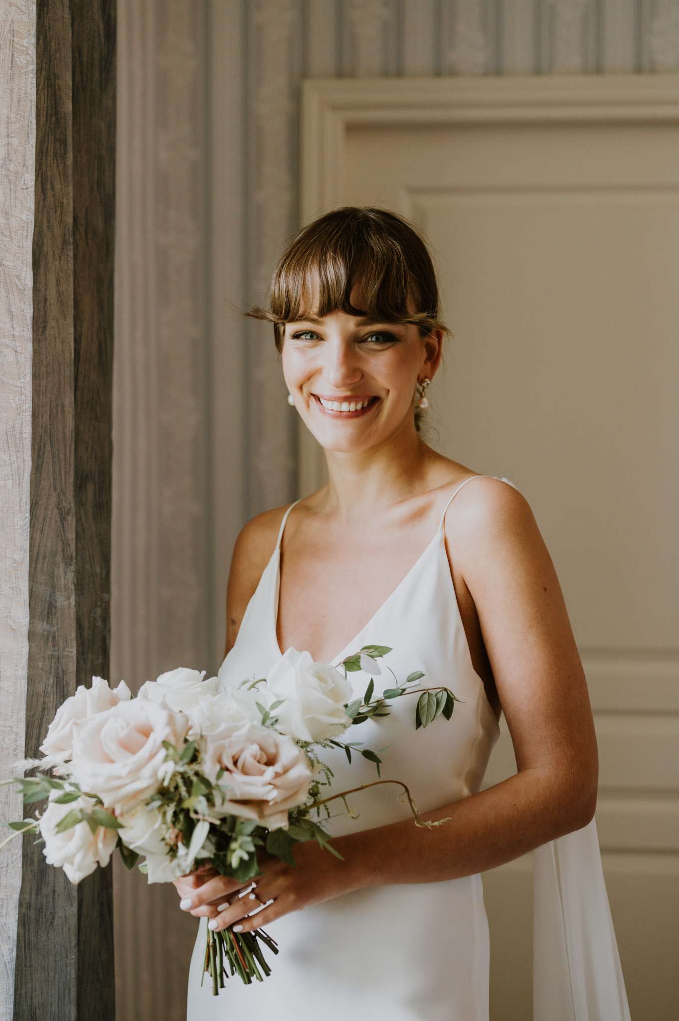 A bridal portrait taken indoors, likely in a château or manor interior, with panelled walls in a warm off-white tone and a linen curtain visible to the left. The bride stands alone, smiling directly at the camera, wearing a minimalist ivory slip-style gown with thin spaghetti straps and a V-neckline. Her dark brown hair is worn up with a short fringe, and she accessorises with pearl drop earrings and a ring. She holds a loosely arranged bouquet of blush and white roses with trailing green foliage. The overall styling is clean and modern with a restrained, paired-back aesthetic. Medium portrait crop, shot from approximately waist height.