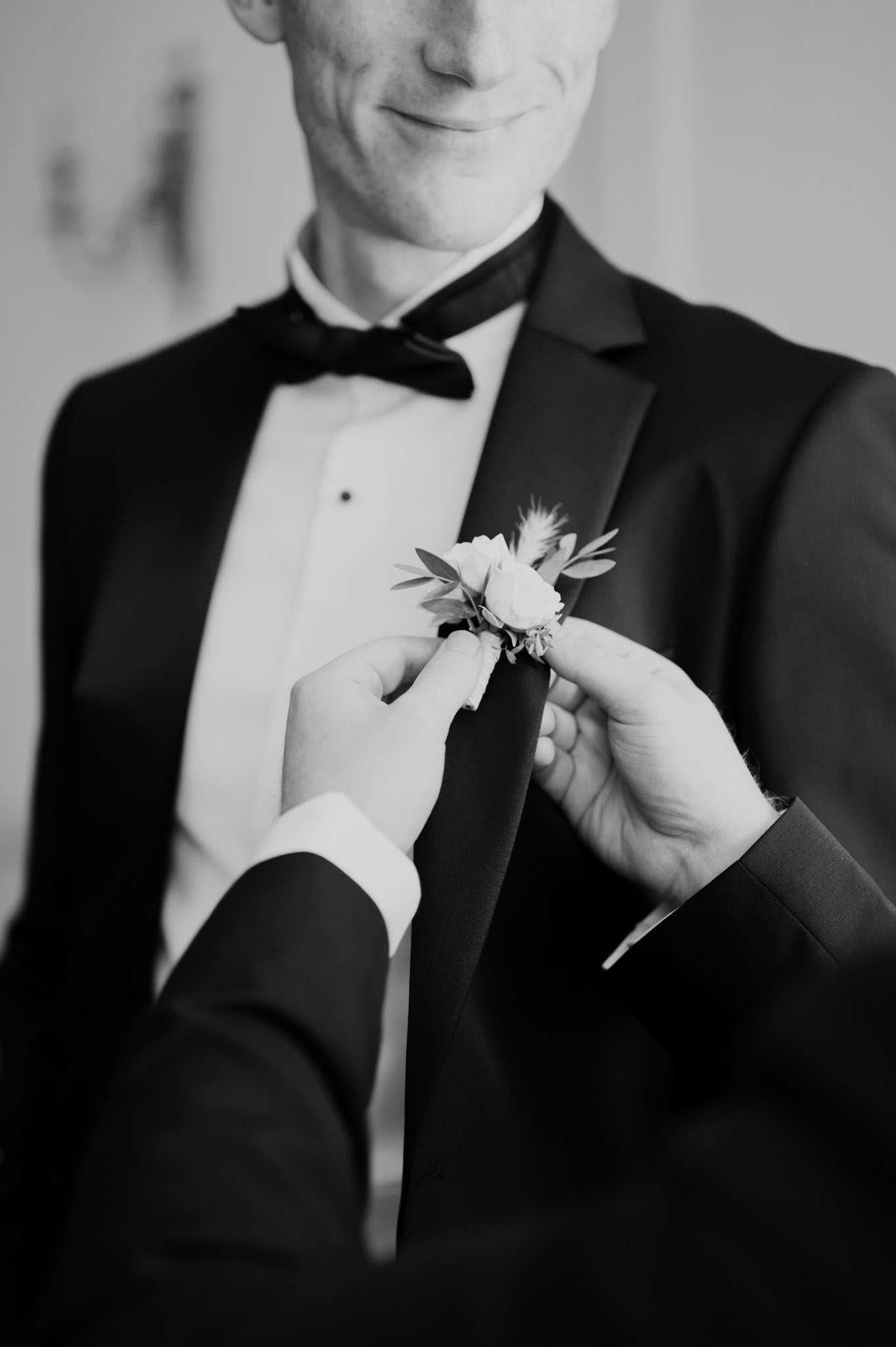 Black and white close-up of hands pinning rose boutonniere on groom's black satin-lapel tuxedo