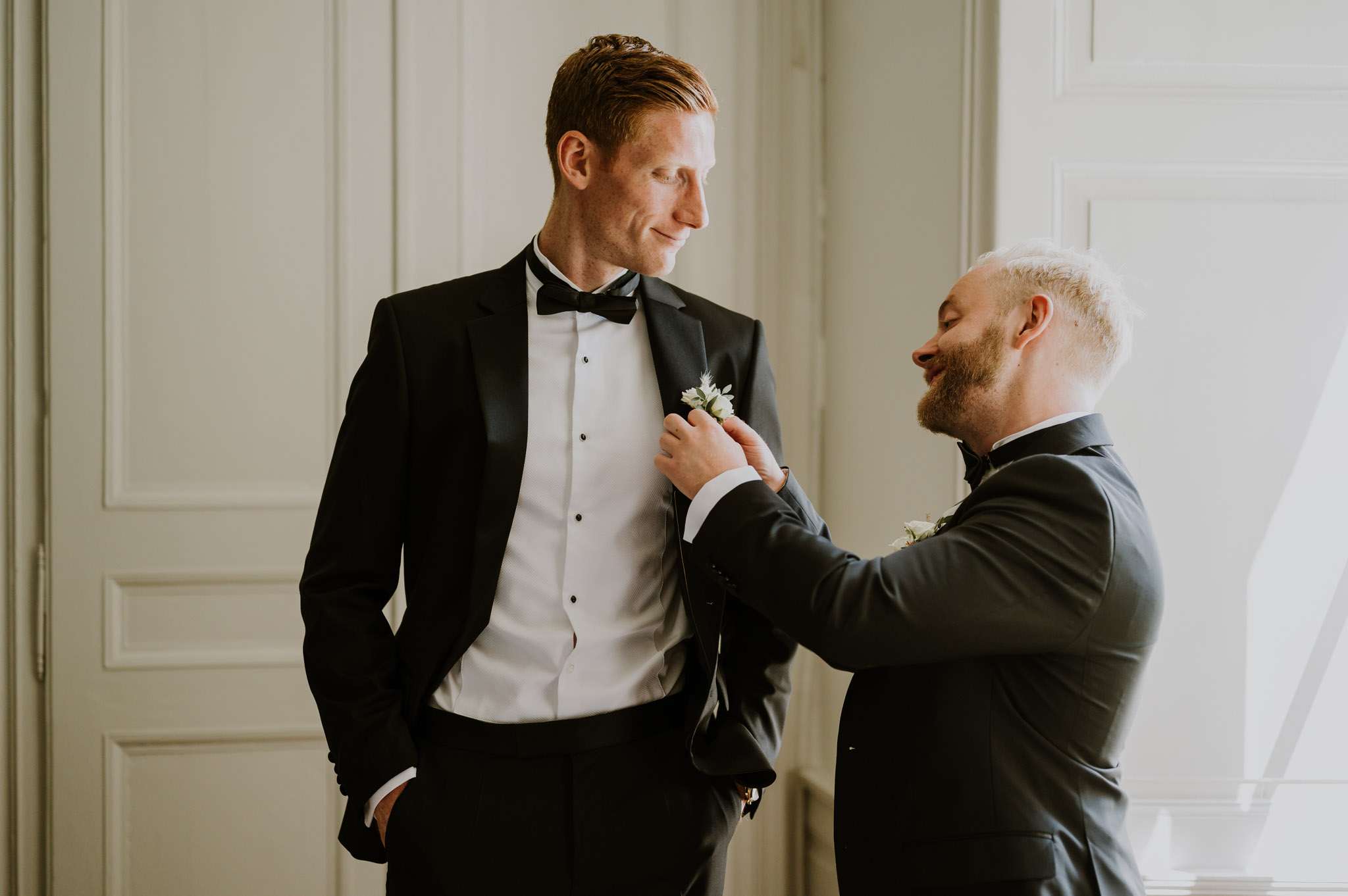 A getting-ready moment captured indoors, showing two men in formal attire in front of white panelled doors. The taller man, dressed in a black tuxedo with a white dress shirt and black bow tie, stands with one hand in his pocket while the second man pins a small white boutonniere with greenery to his lapel. The second man wears a dark charcoal suit with a black bow tie and his own boutonniere, and is smiling as he adjusts the floral detail. The setting appears to be a formal interior room with soft natural light coming from a window to the right. The shot is a medium portrait with a clean, modern-classic styling aesthetic.