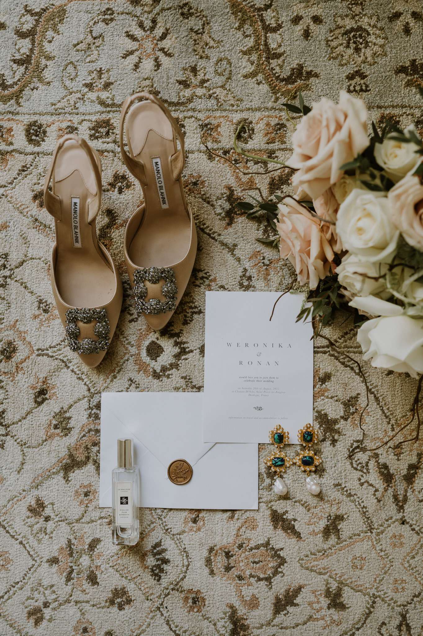 Flat lay of Manolo Blahnik Hangisi heels, wedding invitation with gold seal, Jo Malone perfume and emerald earrings