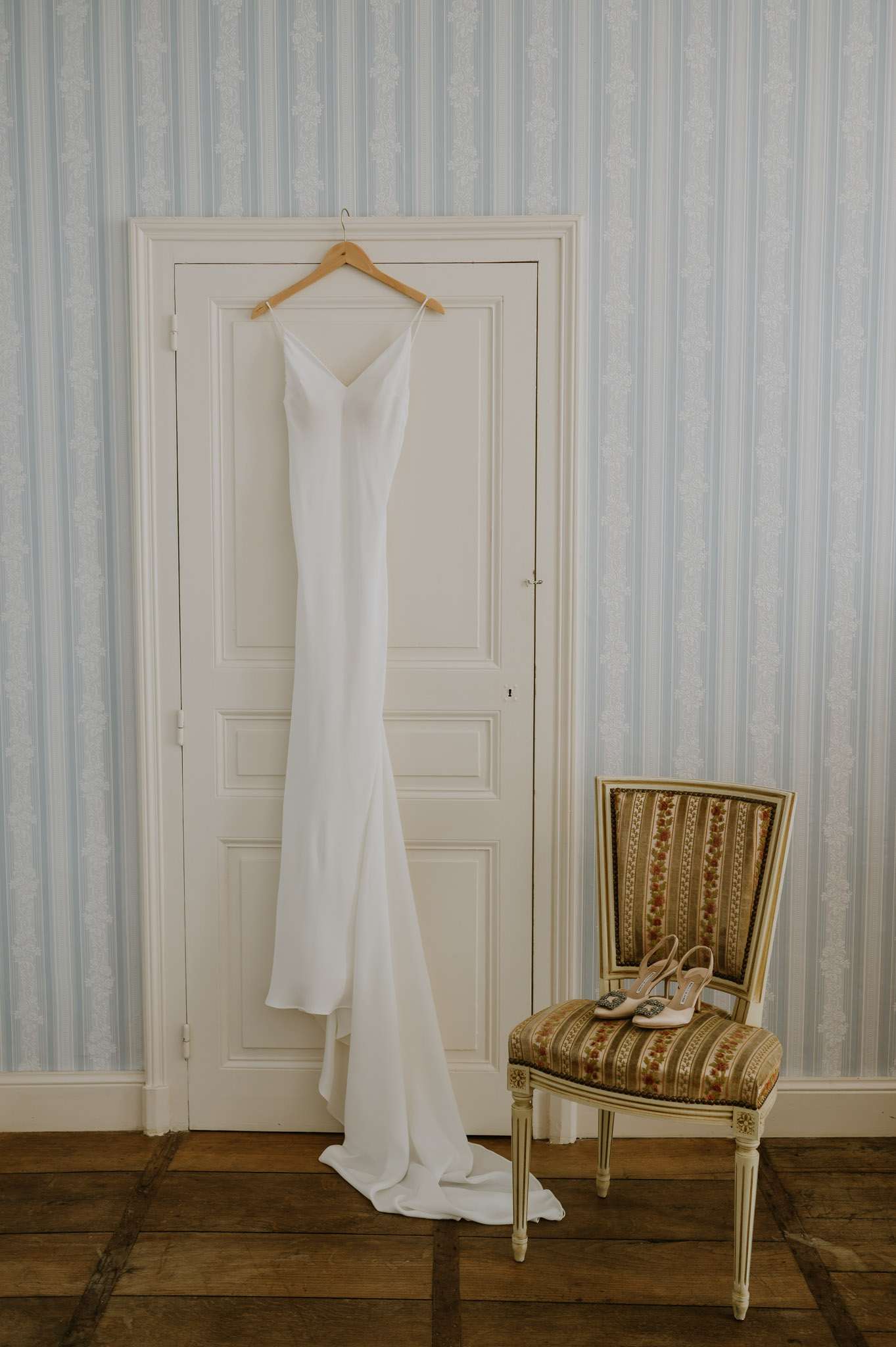 White slip wedding dress on hanger with ivory satin bridal shoes on Louis XVI chair in blue-striped chateau bedroom