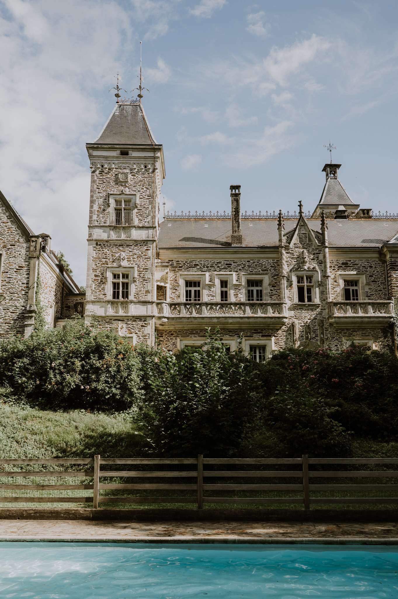 Grey-stone chateau with square tower and pointed slate roof viewed from turquoise pool across sloped garden